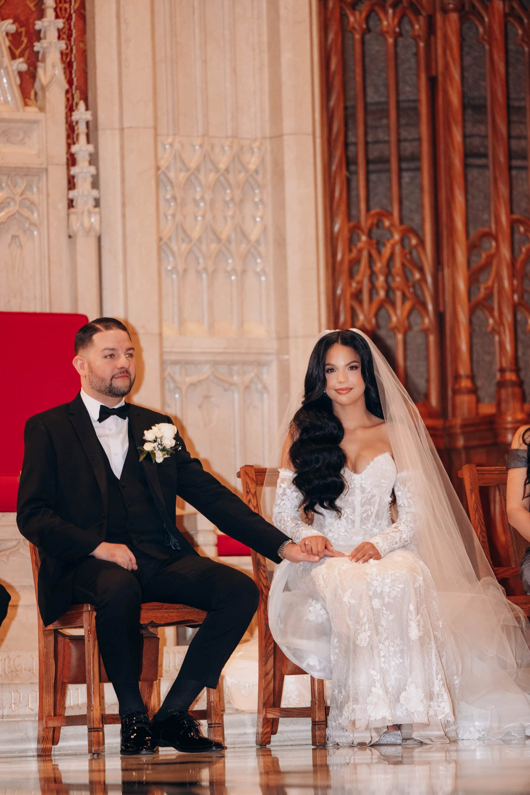Bride in lace gown and veil holding hands with groom in black tuxedo seated inside The Cathedral Basilica of the Sacred Heart with ornate stone and wood background.