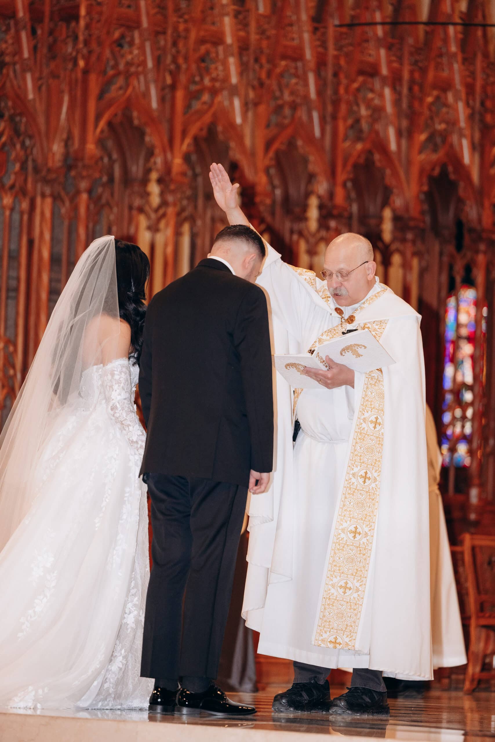 Bride and groom receiving a blessing from a priest in a richly detailed cathedral with carved wooden arches and stained glass windows.