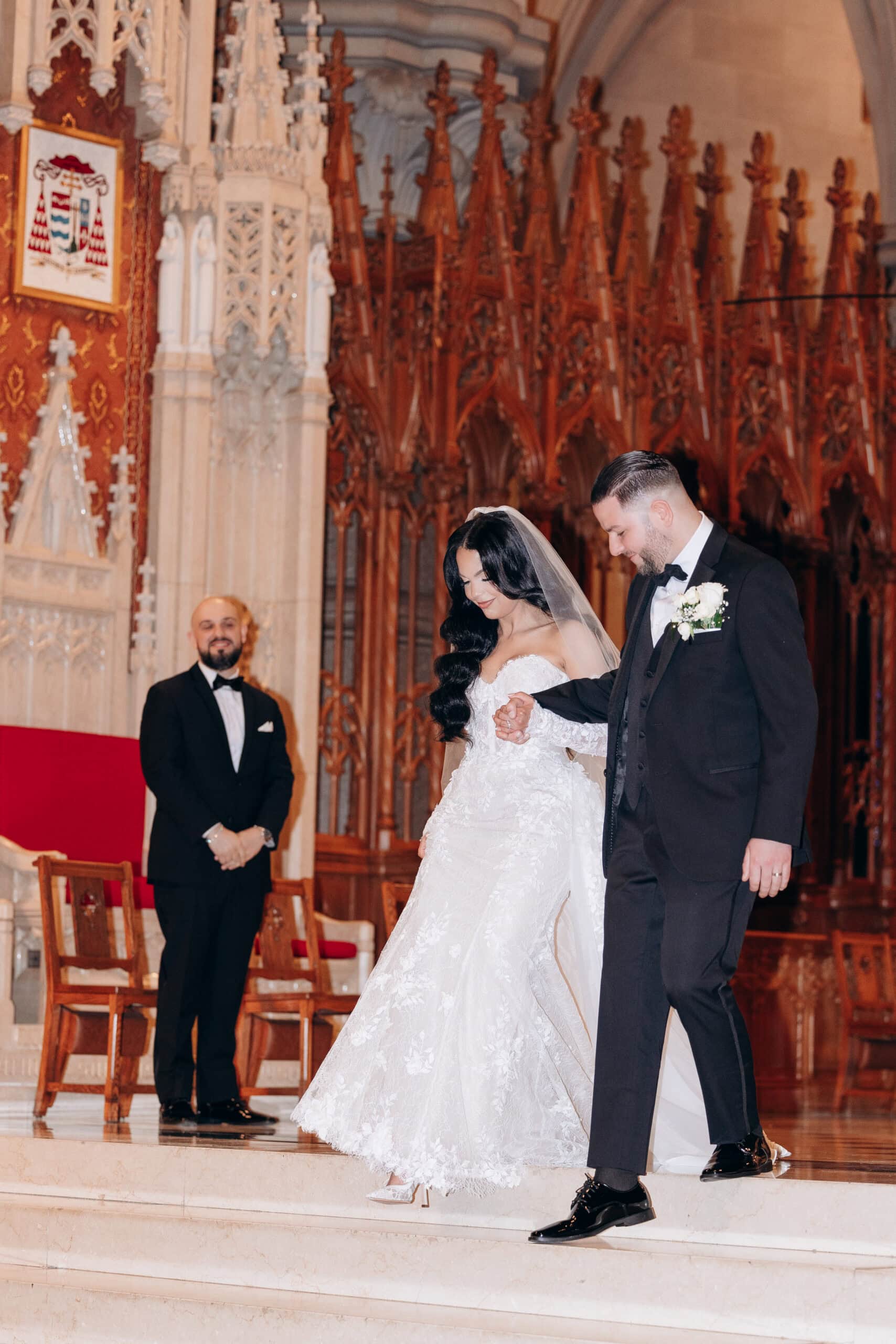Bride in detailed lace gown and veil, holding hands with groom in black tuxedo, descending marble steps inside The Cathedral Basilica of the Sacred Heart with ornate wooden backdrop.