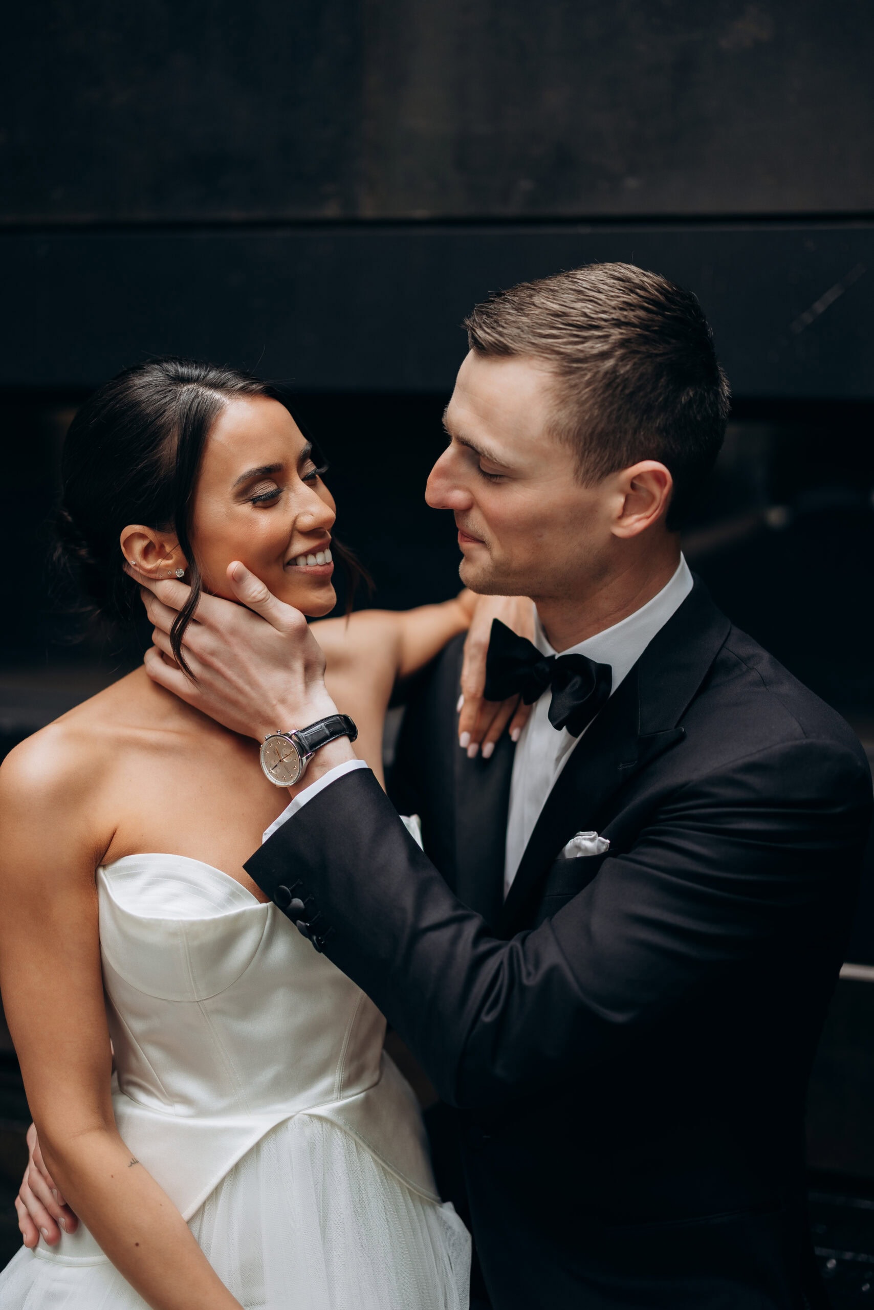 Bride in a strapless satin gown and groom in a black tuxedo share a close portrait at The Foundry, with the groom gently holding her face against a dark backdrop.