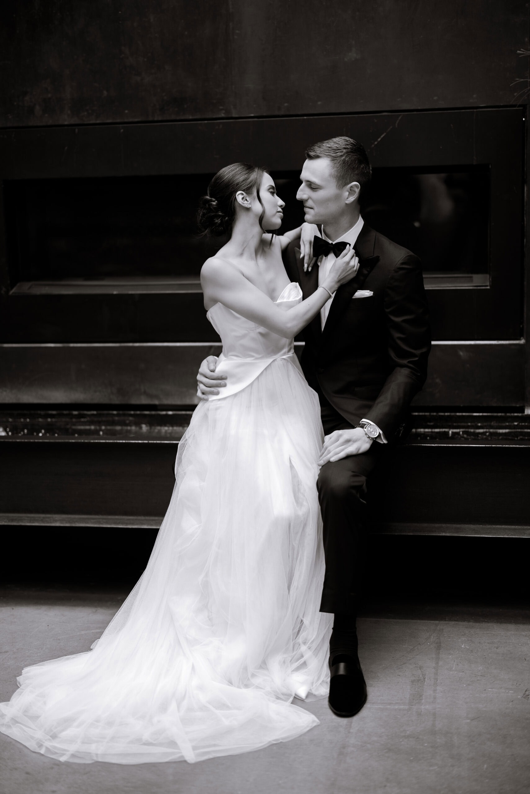 Black-and-white wedding portrait at The Foundry of a bride in a strapless tulle gown adjusting the groom’s bow tie as they sit closely before a dark fireplace.