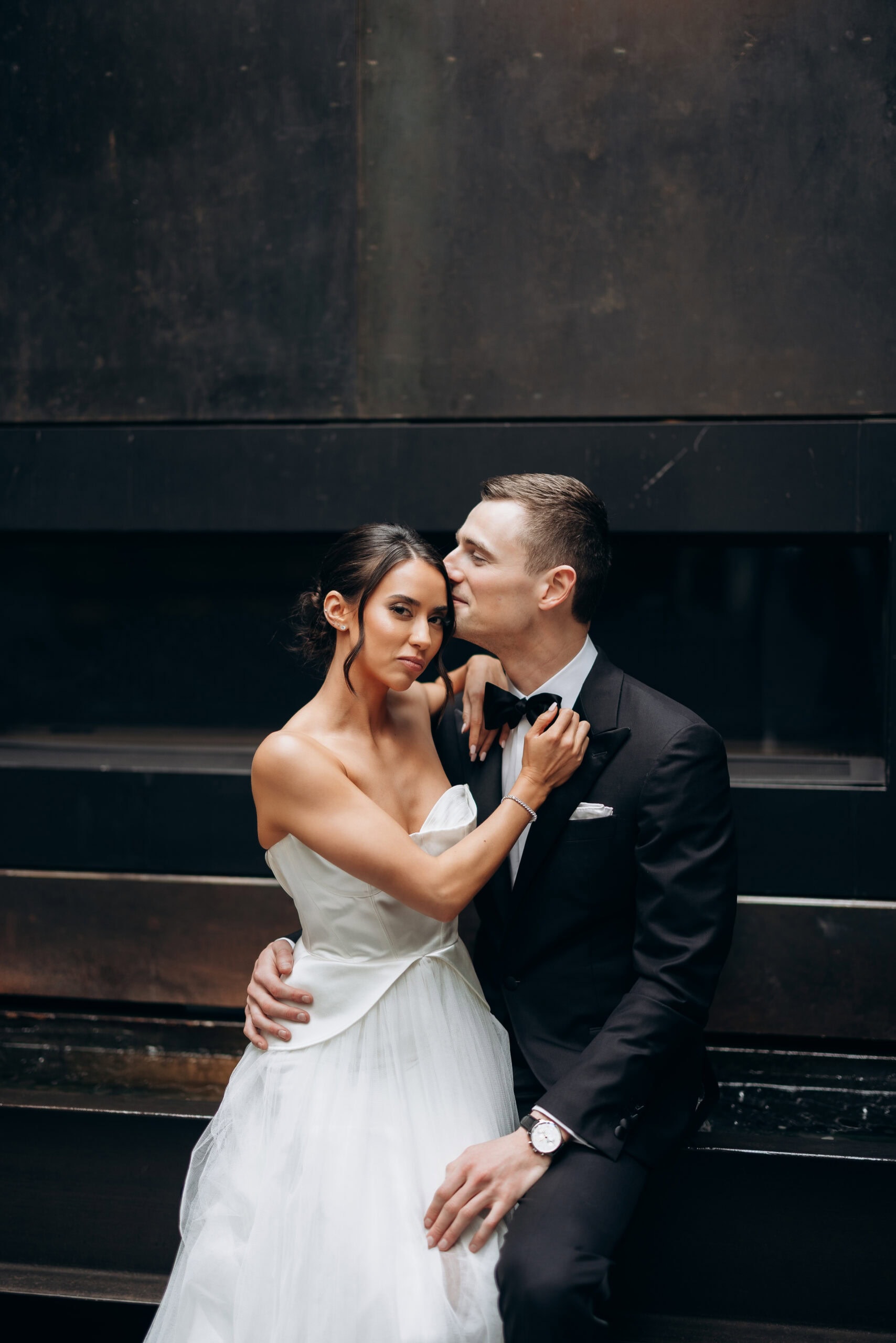 Bride in a strapless satin gown and groom in a black tuxedo pose closely against a dark modern wall at The Foundry during a wedding portrait.