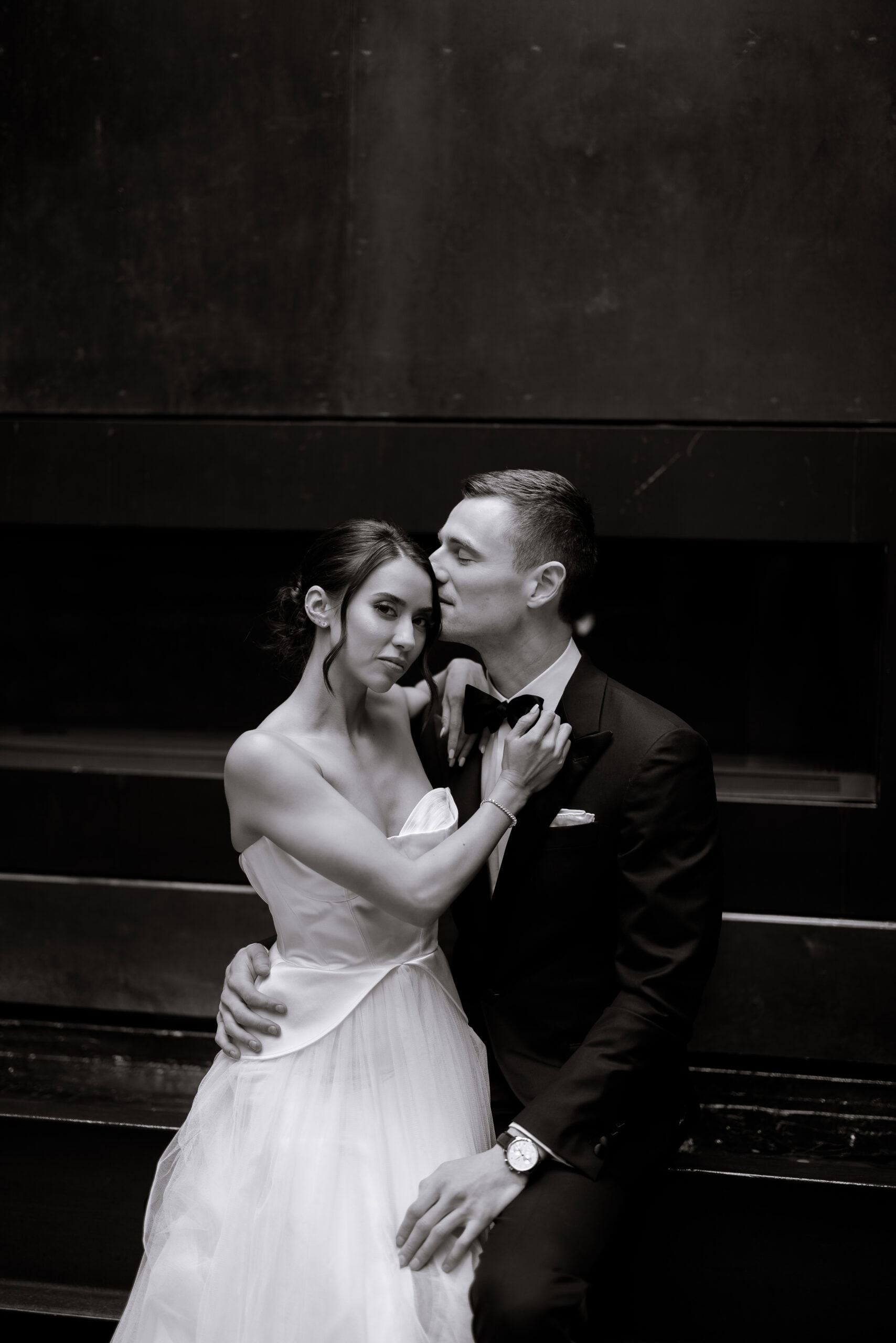 Black-and-white wedding portrait at The Foundry of a bride in a strapless gown embracing a groom in a tuxedo as he kisses her temple against a dark wall.