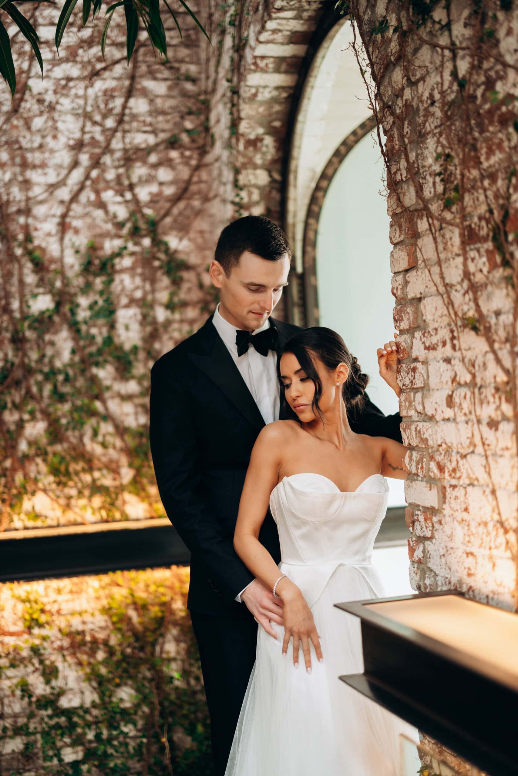 Bride in a strapless white gown and groom in a black tuxedo standing closely beneath a brick arch with vines at The Foundry during a wedding portrait.