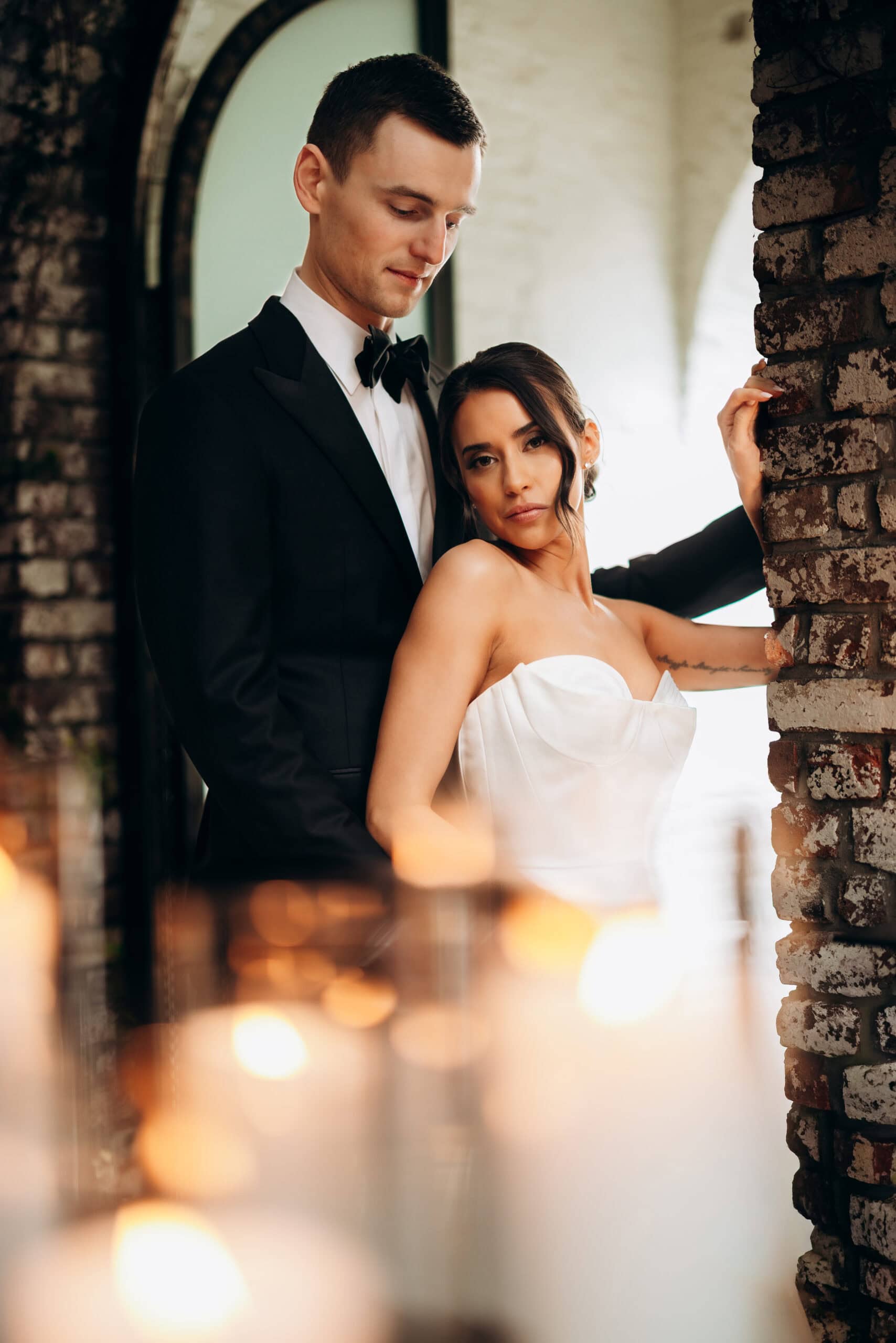 Bride in a strapless satin gown and groom in a black tuxedo pose beneath brick arches at The Foundry, with soft candlelight blurred in the foreground.