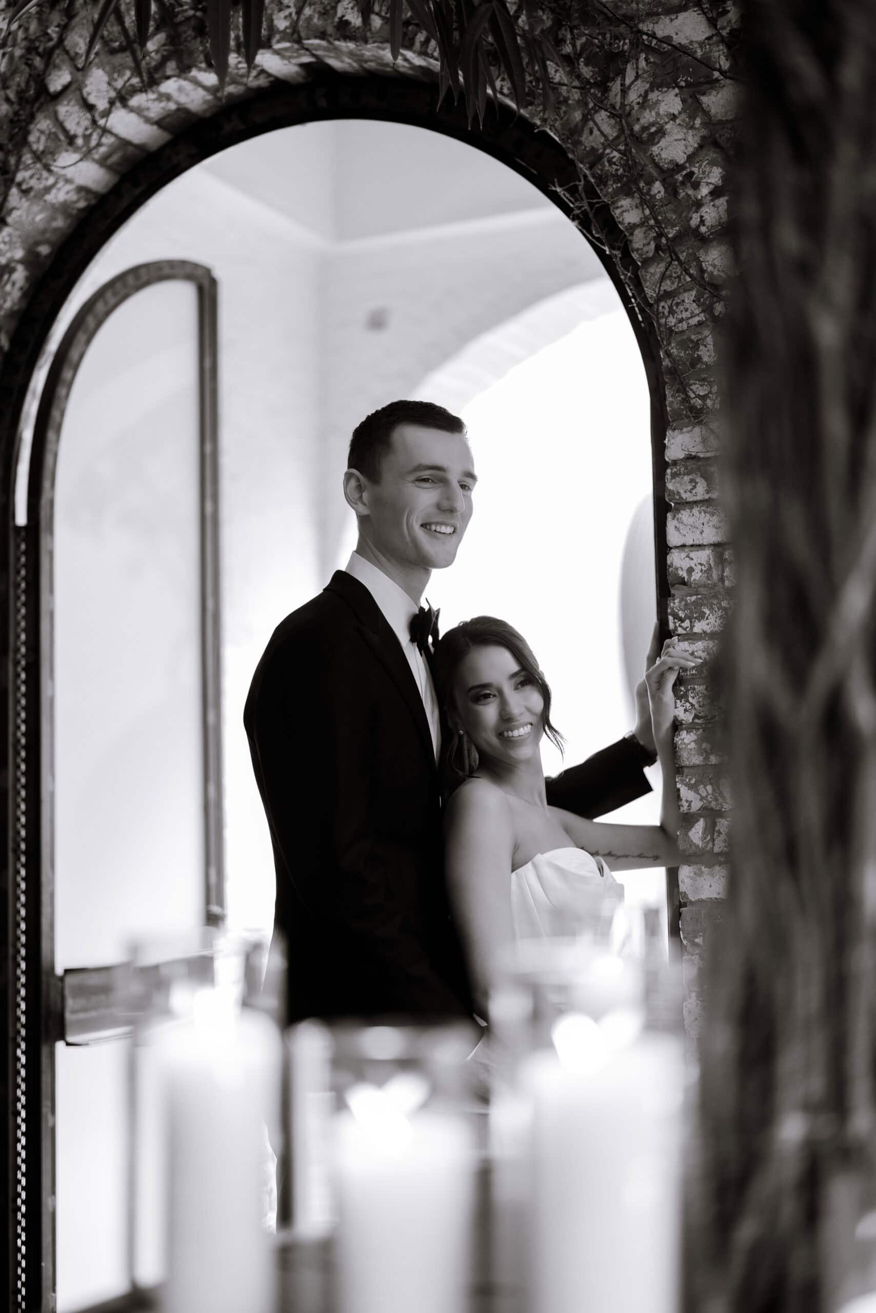 Black-and-white wedding portrait of a couple at The Foundry, standing beneath a rustic brick arch indoors with blurred candles in the foreground.