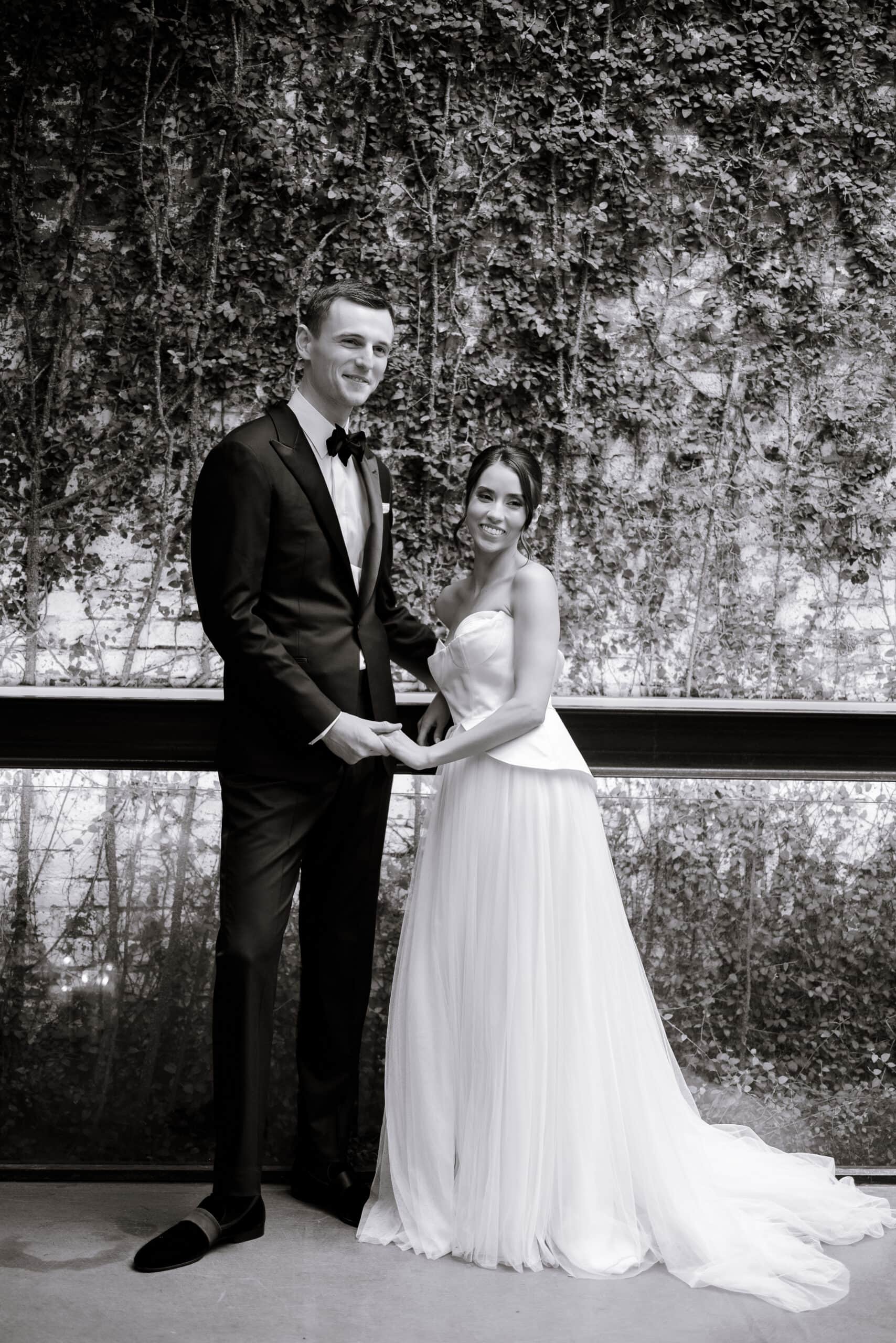 Black-and-white wedding portrait at The Foundry of a couple holding hands before an ivy-covered wall; he wears a tuxedo, she wears a strapless gown with a full skirt.
