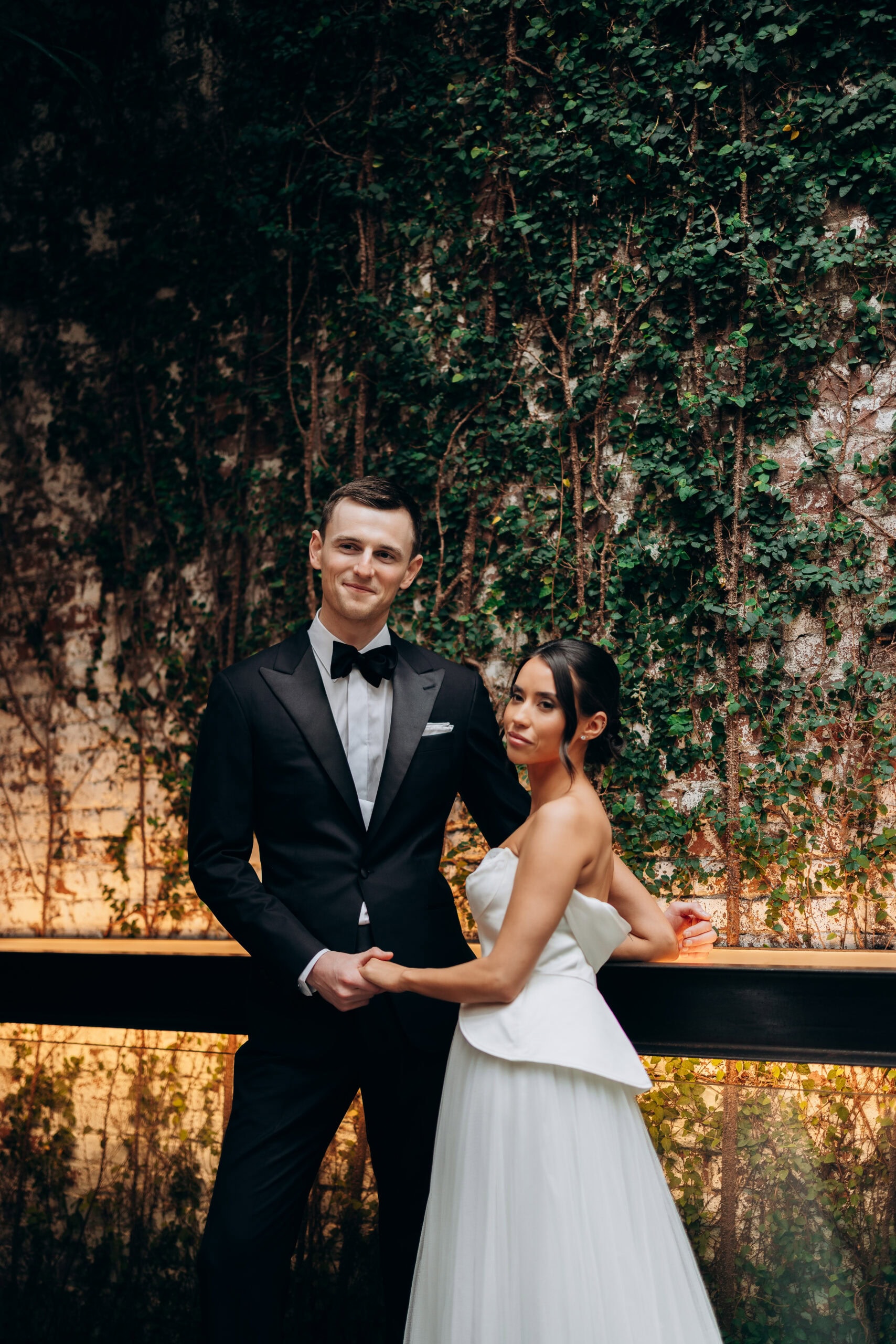 Bride and groom pose at The Foundry before an ivy-covered wall, with warm uplighting, a black tuxedo, and a strapless white gown in an editorial portrait.