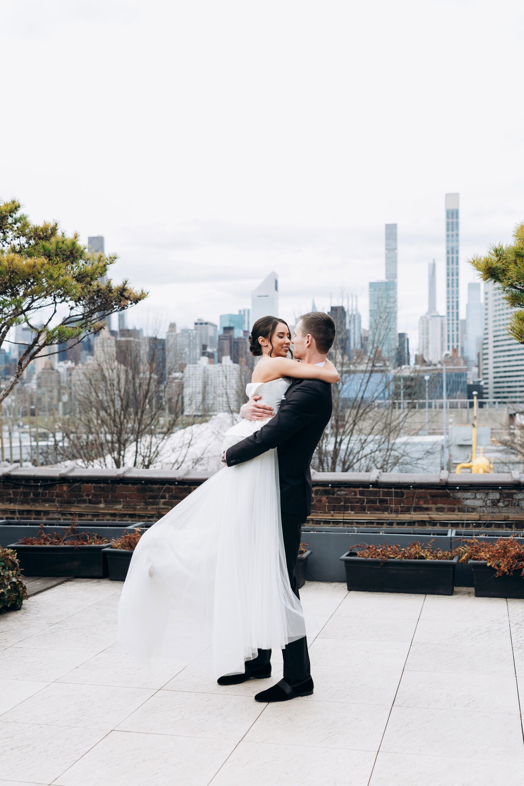 Bride in a strapless tulle gown embraces groom in a black suit on a rooftop terrace at The Foundry, with the Manhattan skyline behind them on an overcast day.
