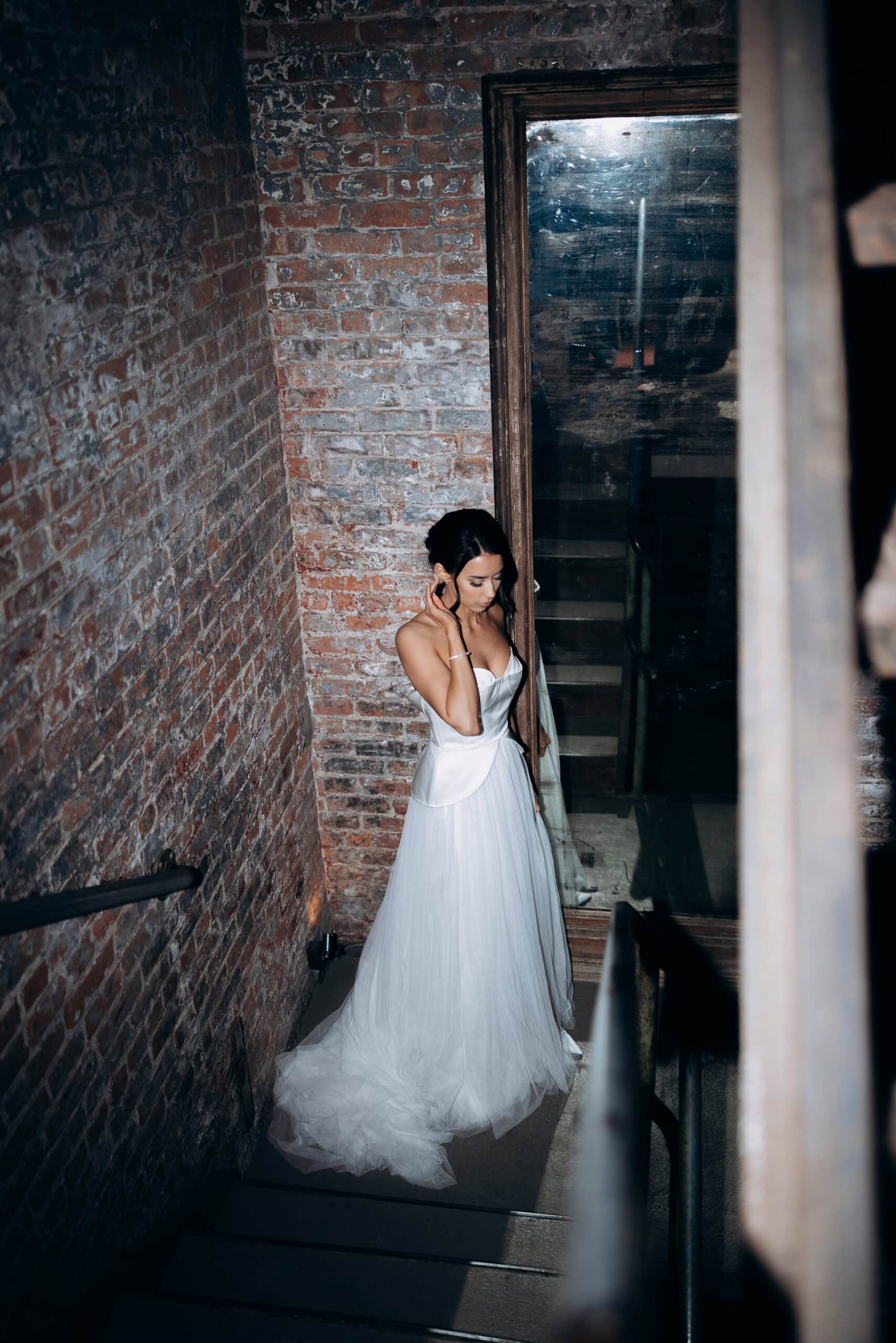 Bride in a strapless white gown standing on a narrow stairwell at The Foundry, surrounded by exposed brick walls and a large aged mirror.