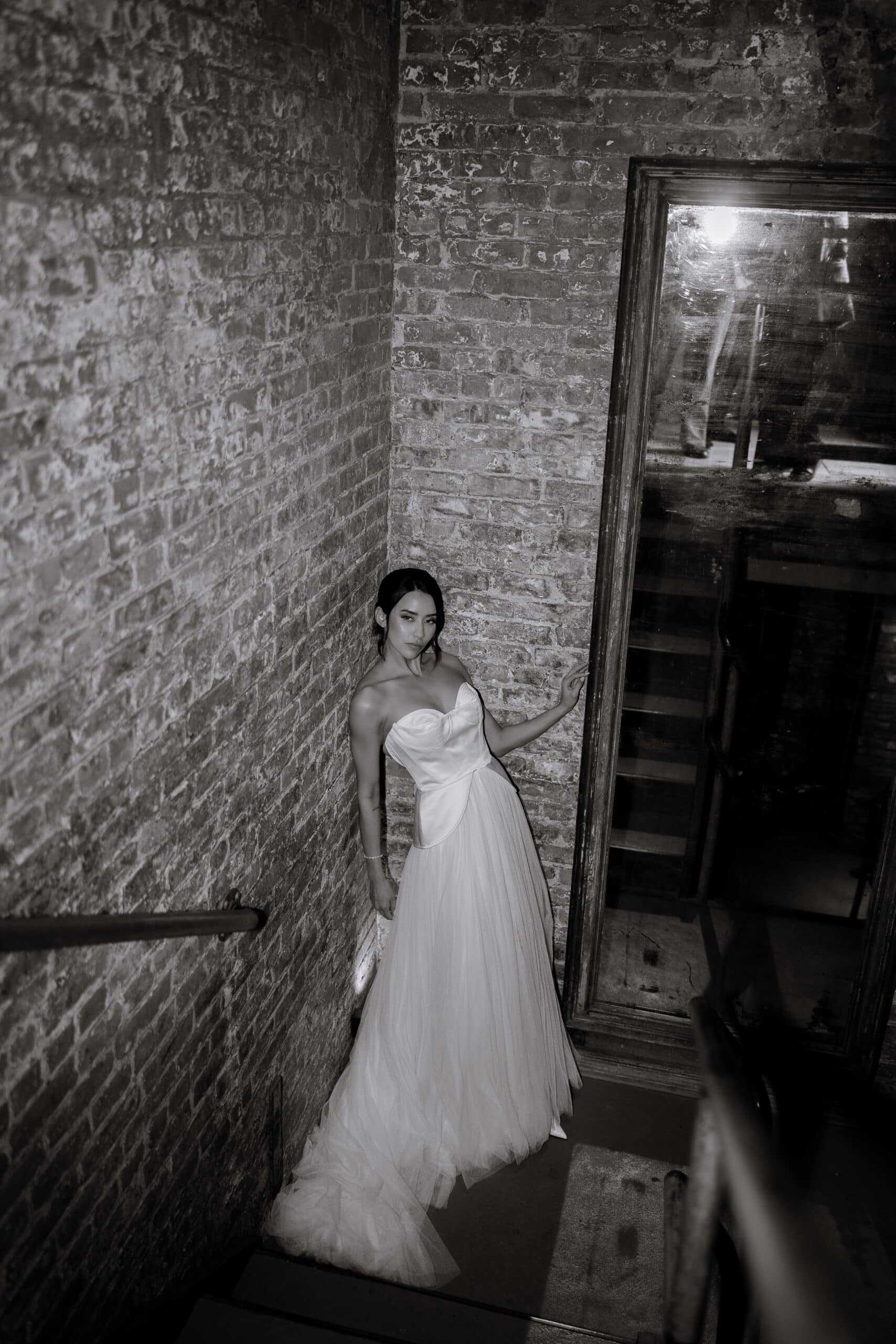 Black-and-white bridal portrait at The Foundry showing a woman in a strapless satin gown standing on a narrow brick stairwell beside a large vintage mirror.