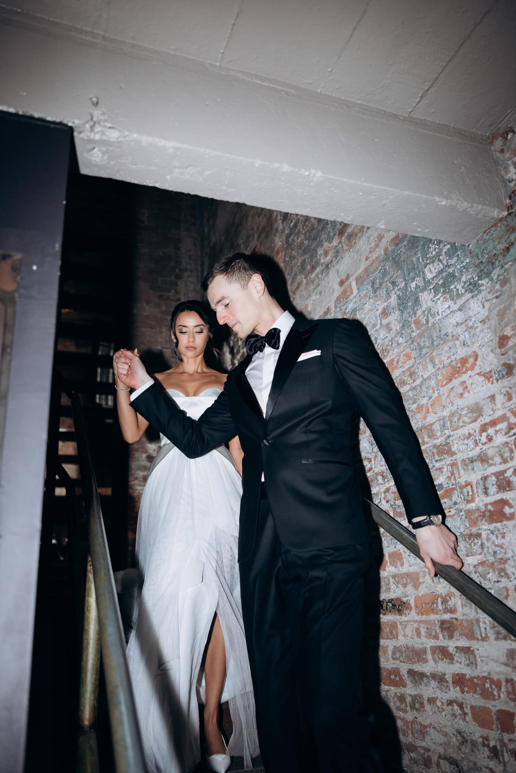 Bride and groom descend a narrow brick stairway at The Foundry, she in a strapless white gown and heels, he in a black tuxedo, holding hands.