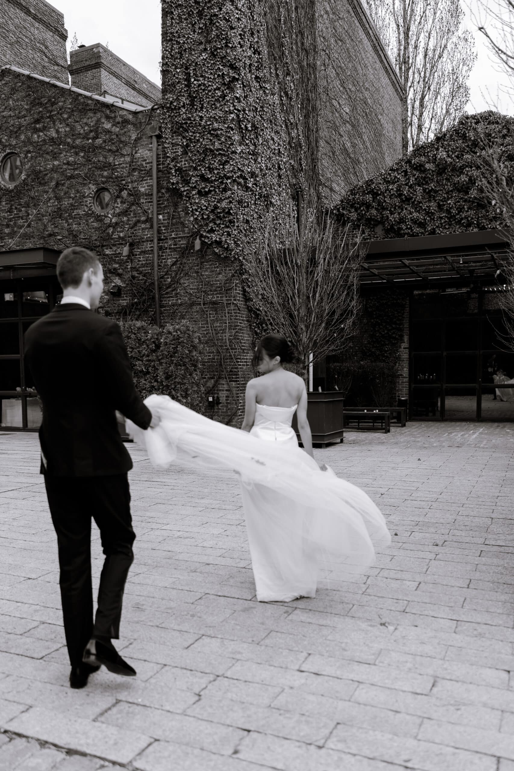 Black-and-white wedding portrait at The Foundry showing a couple from behind in an ivy-covered brick courtyard, with the groom holding the bride’s flowing veil.