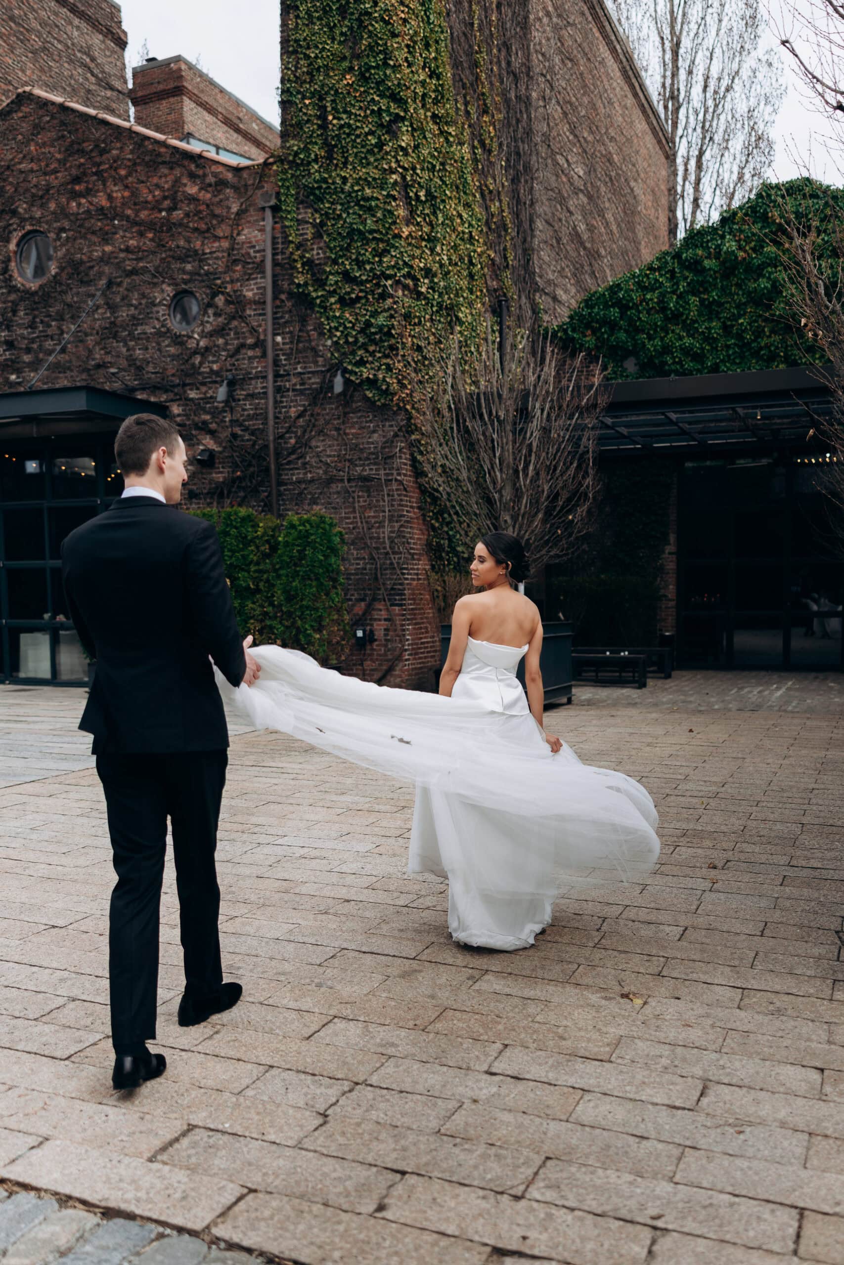 Bride in a strapless gown walks across The Foundry courtyard as her partner holds her long veil, framed by ivy-covered brick walls and a paved terrace.