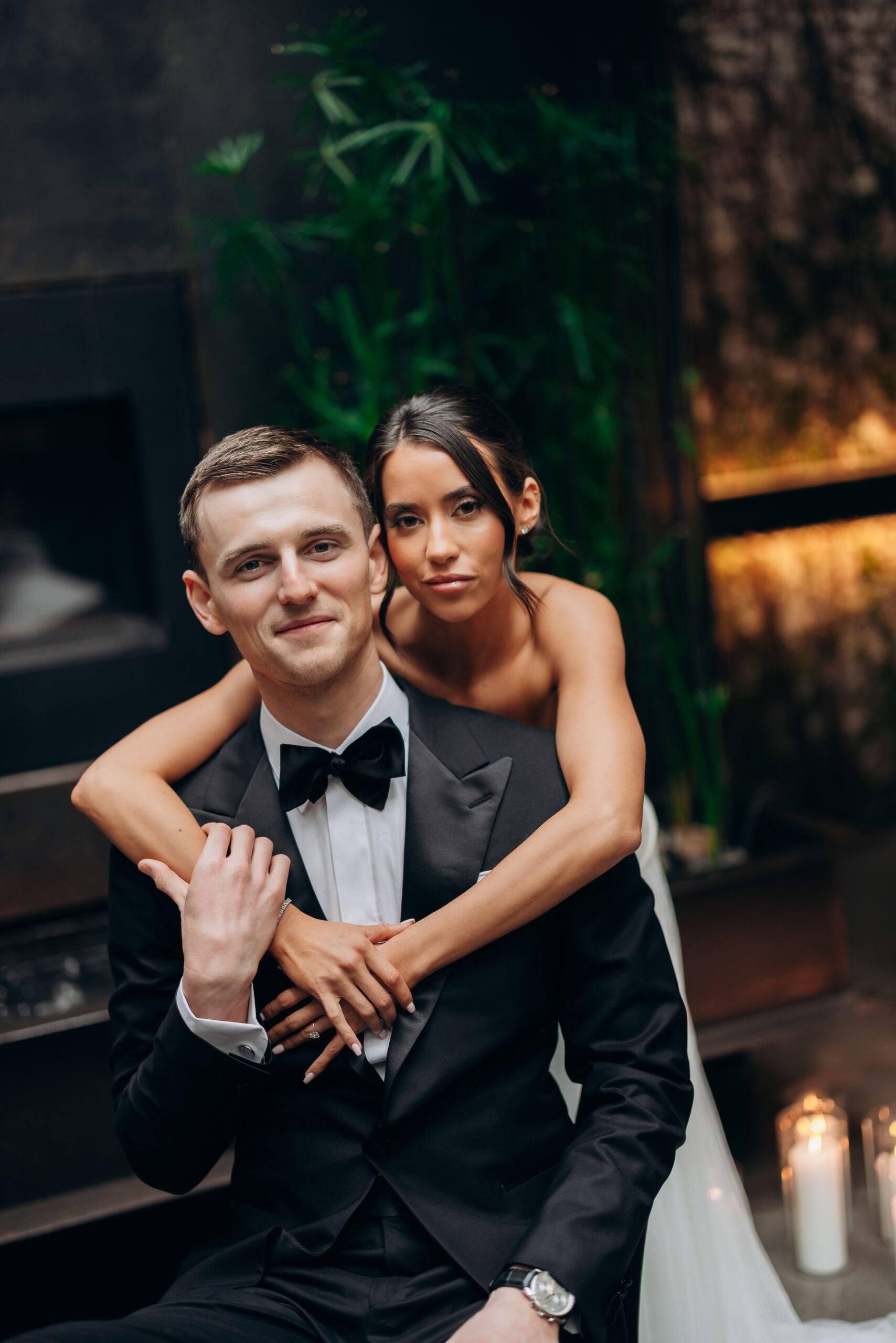 Bride embraces groom from behind in a black-tie wedding portrait indoors at The Foundry, with candles, dark stone, and green plants in the background.