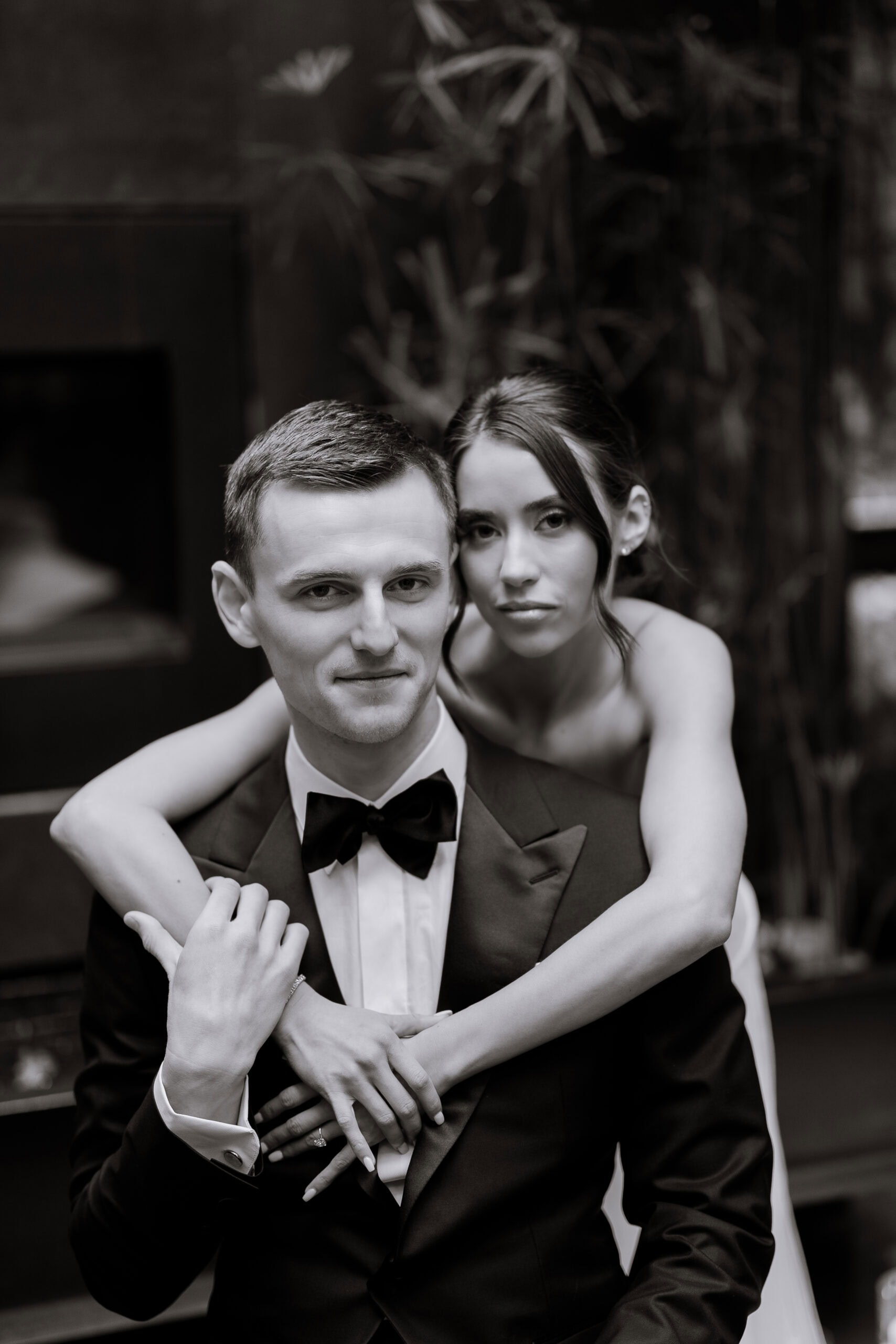 Black-and-white portrait of a couple at The Foundry, with the woman embracing the seated man in a tuxedo against a softly blurred interior backdrop.