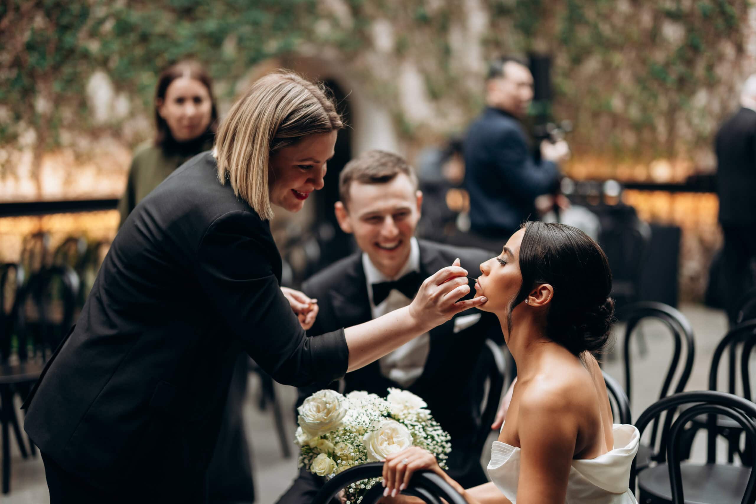 At The Foundry, a makeup artist applies a final touch to a seated bride in a strapless satin gown while a groom in a tuxedo sits beside her holding white roses.