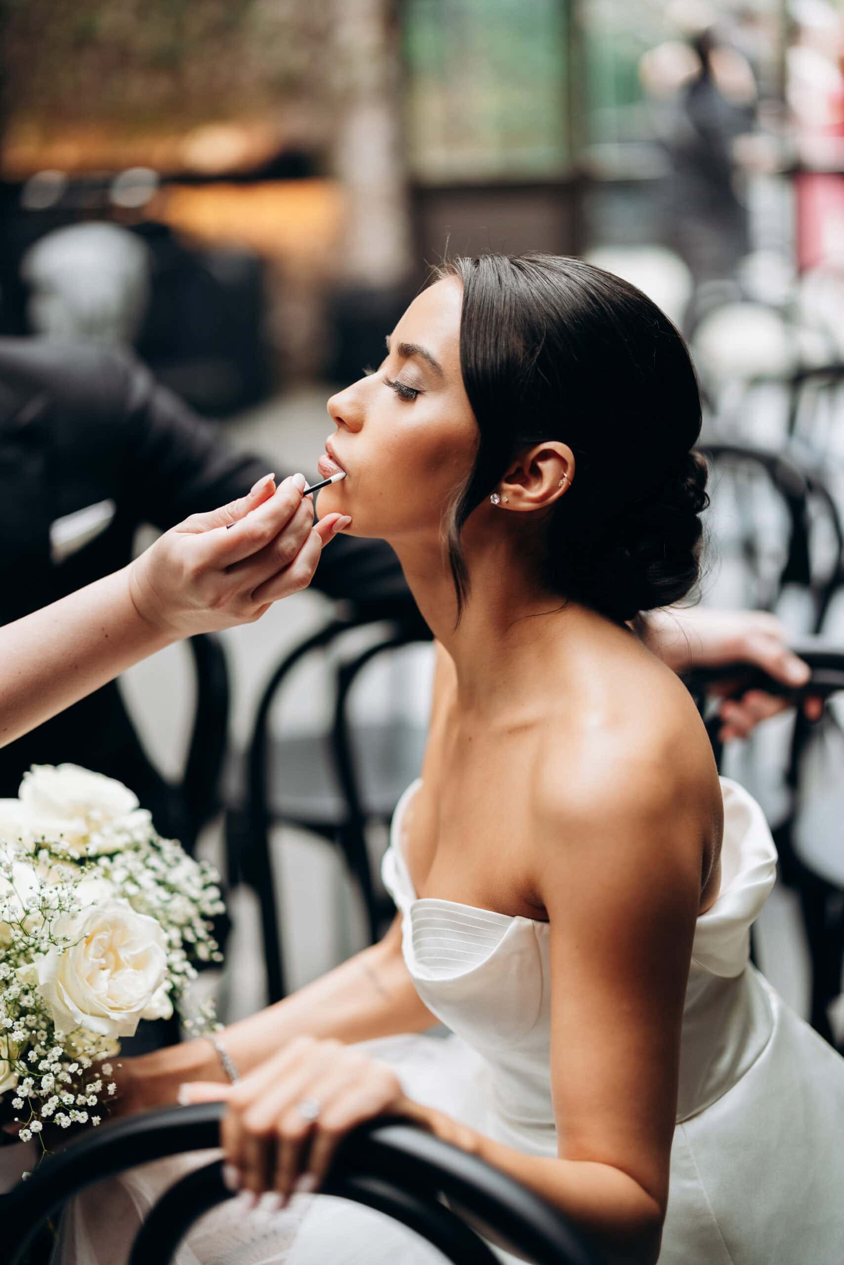 Bride seated indoors at The Foundry while a makeup artist applies lip color; she wears a strapless satin gown and holds a bouquet of white roses and baby's breath.