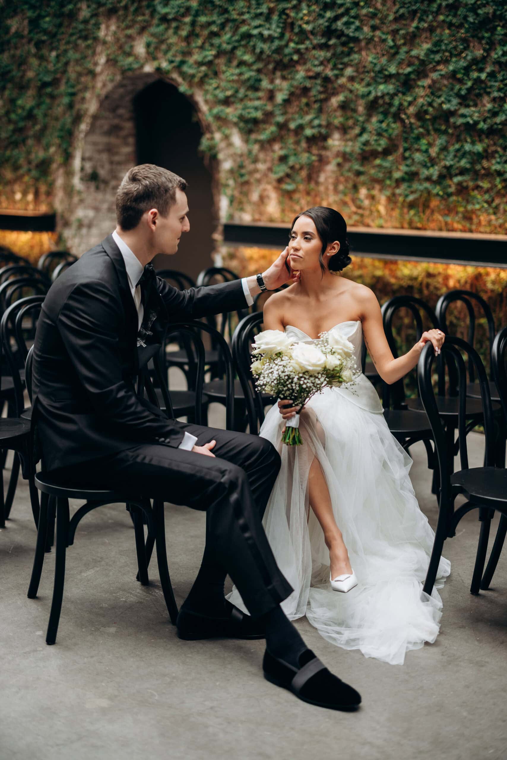 Bride and groom seated among black chairs at The Foundry, with an ivy-covered stone arch behind them; she holds a white rose bouquet in a strapless gown.