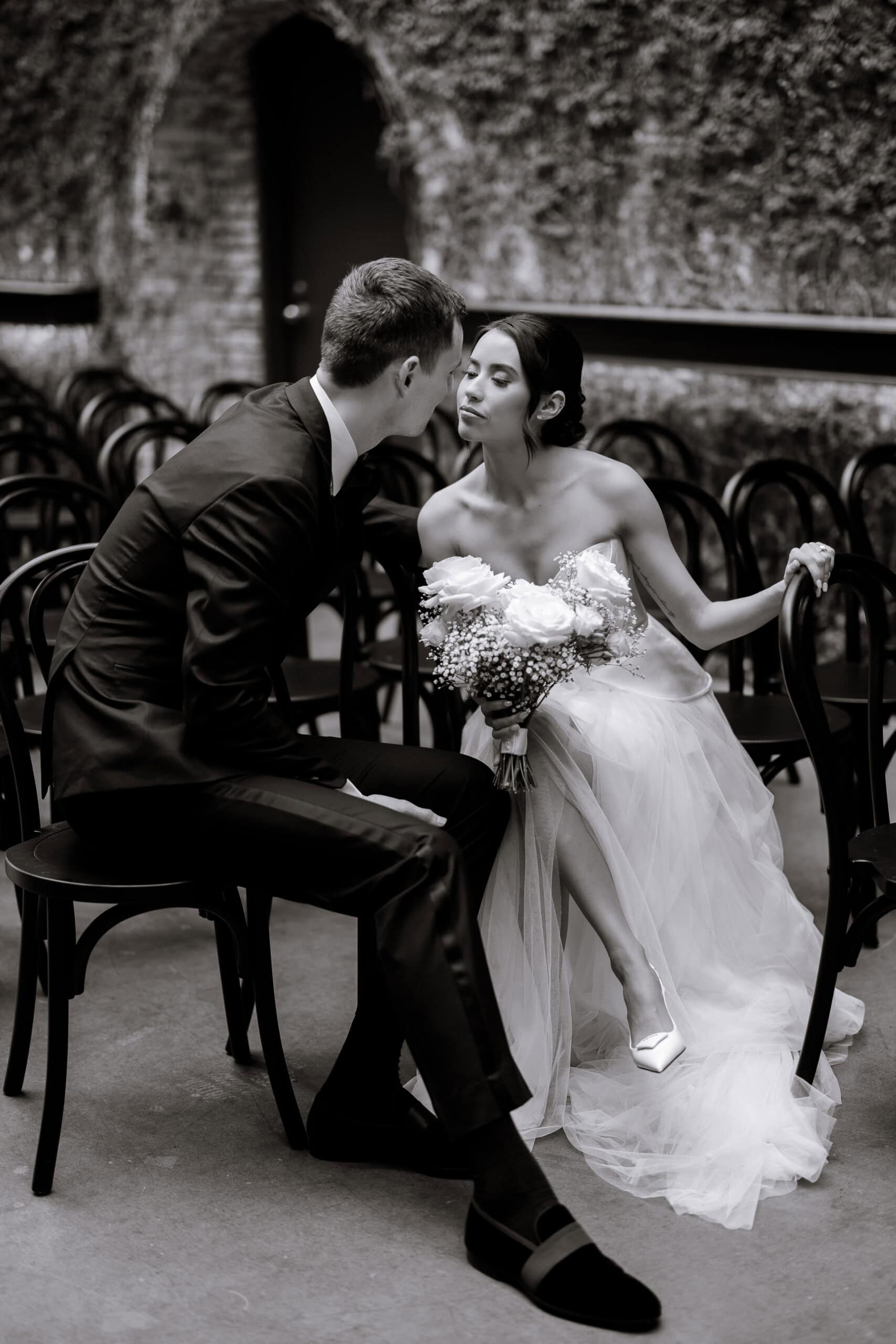 Black-and-white wedding portrait at The Foundry of a seated couple leaning in for a kiss among black chairs; bride in strapless tulle gown holds a rose bouquet.