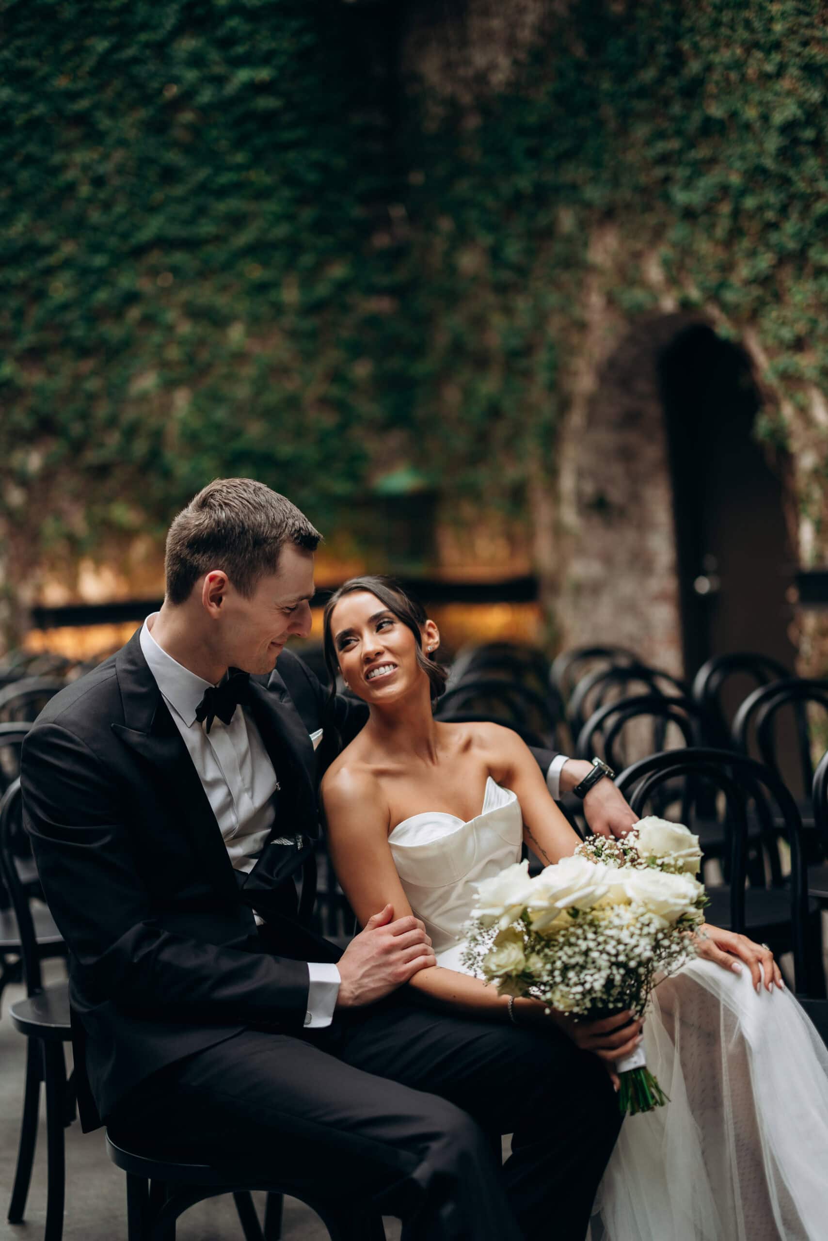 Bride and groom seated together after the ceremony at The Foundry, surrounded by black chairs and an ivy-covered stone wall, holding a white rose bouquet.