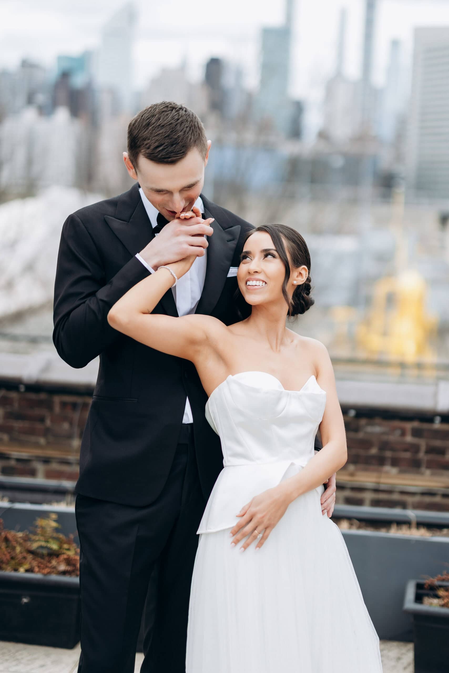 Bride and groom on a rooftop at The Foundry, with the groom kissing her hand and a blurred Manhattan skyline behind them.