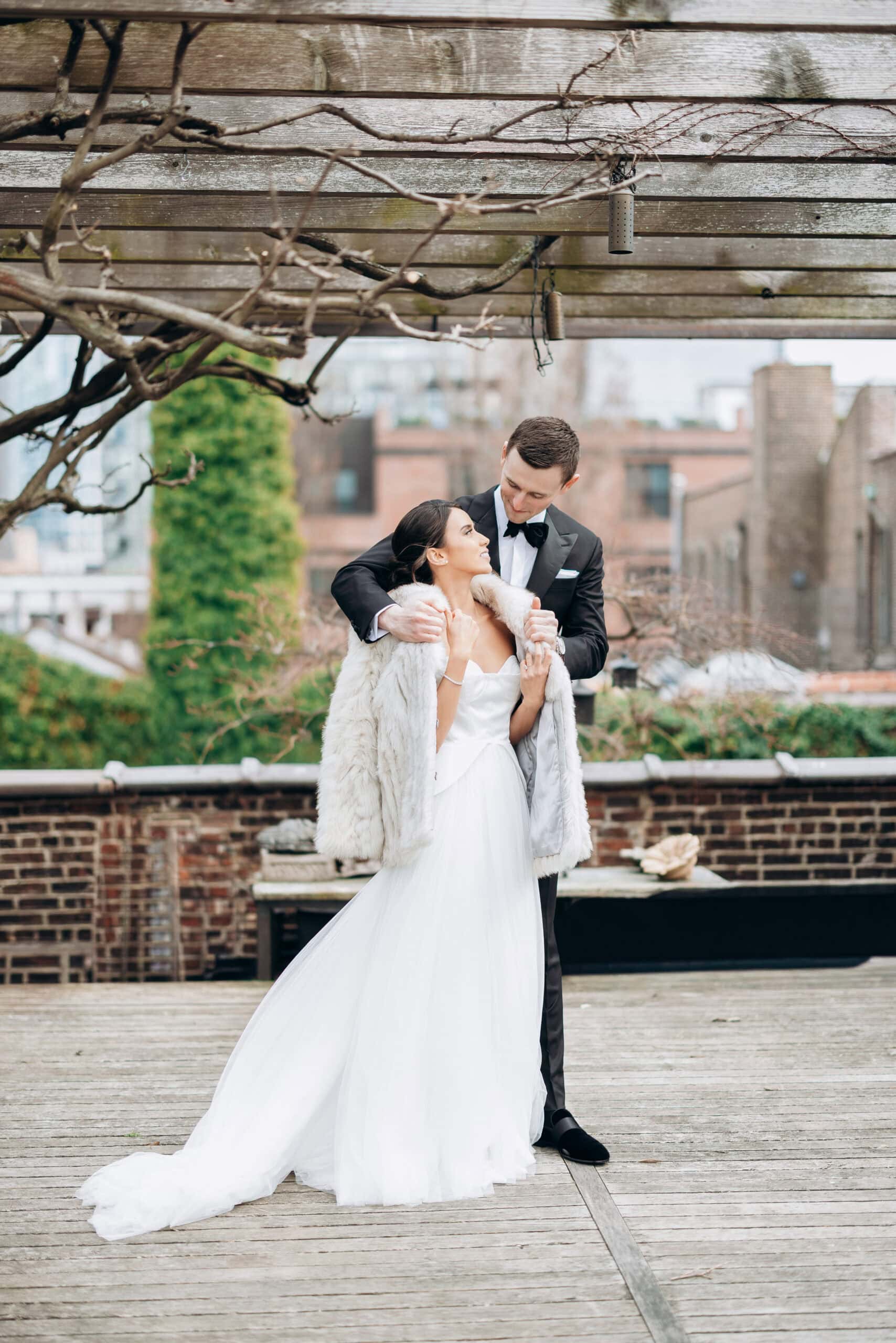 Bride in a white gown and fur wrap stands with groom in a black tuxedo under a wooden pergola on the terrace at The Foundry, with city buildings beyond.