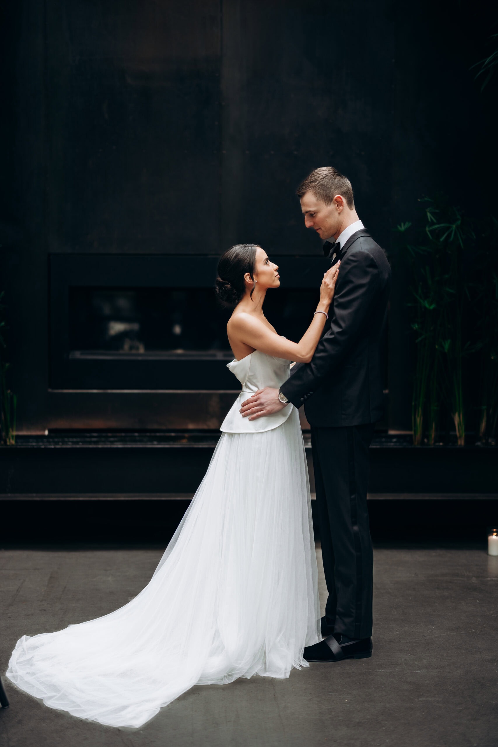 Bride in a strapless white gown and groom in a black tuxedo standing closely indoors at The Foundry before a dark fireplace, facing each other in a formal portrait.