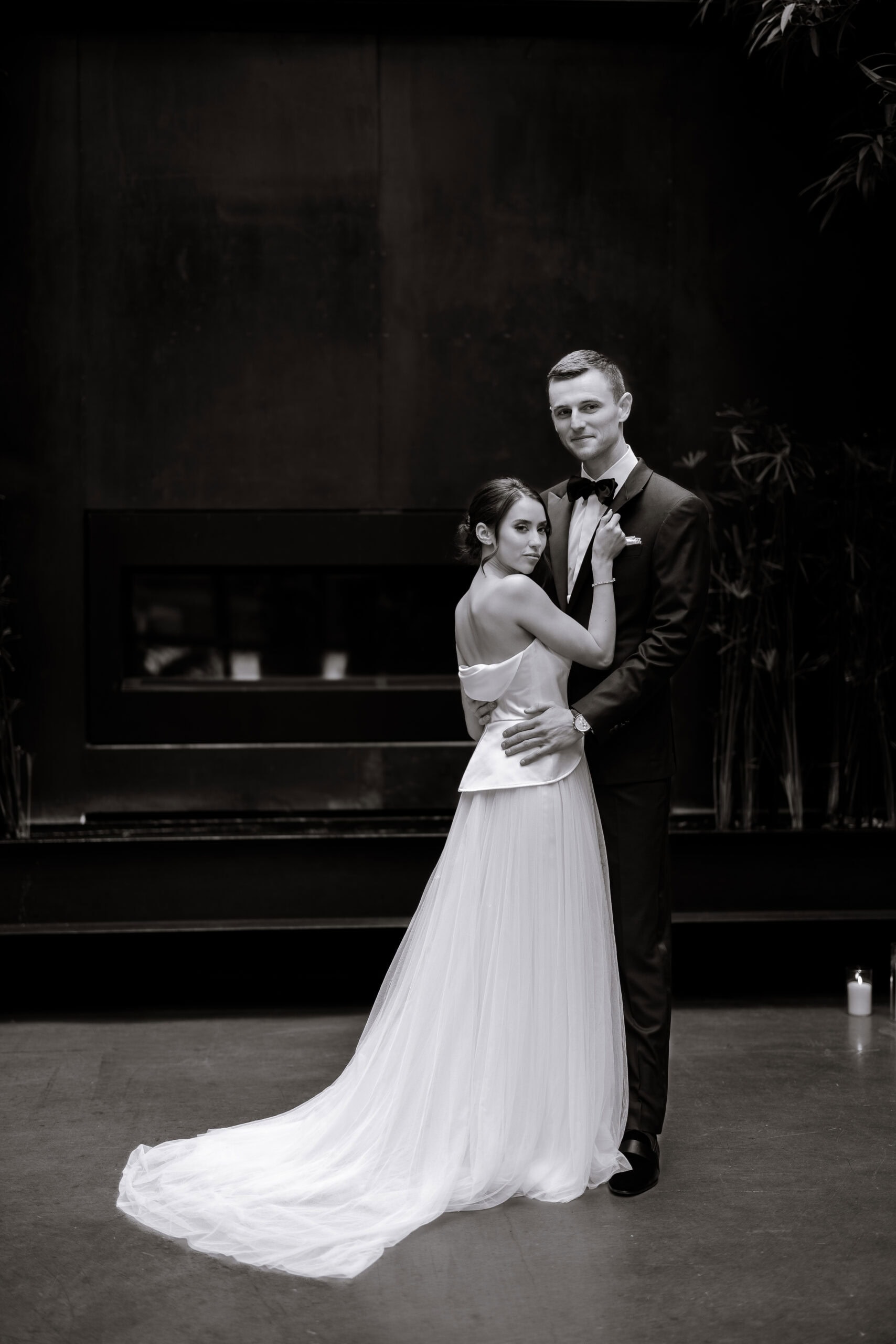 Black-and-white wedding portrait at The Foundry of a couple embracing indoors, with the bride in a low-back tulle gown and the groom in a tuxedo before a dark fireplace.