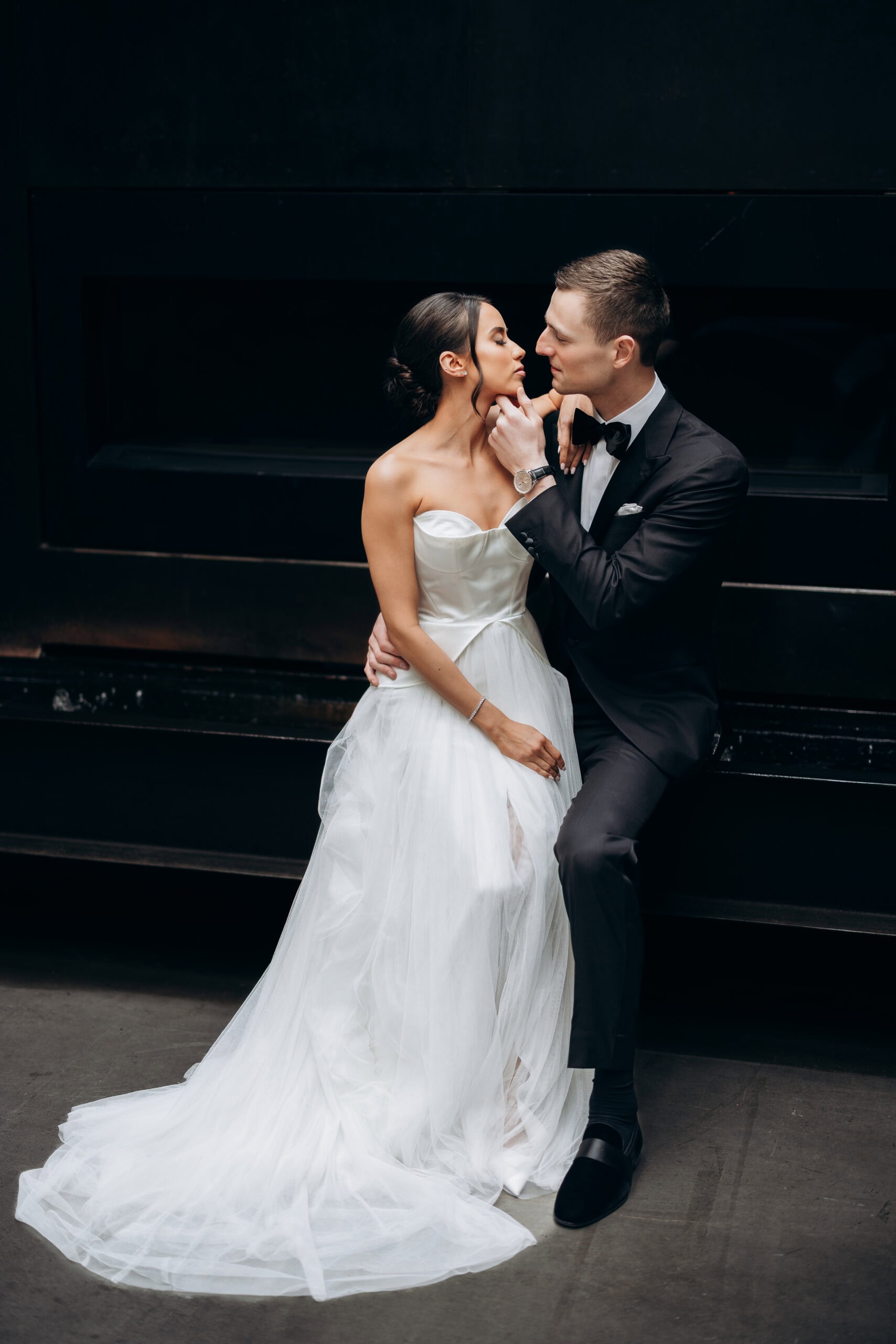 Bride in a strapless white gown and groom in a black tuxedo share an intimate portrait against a dark wall at The Foundry.