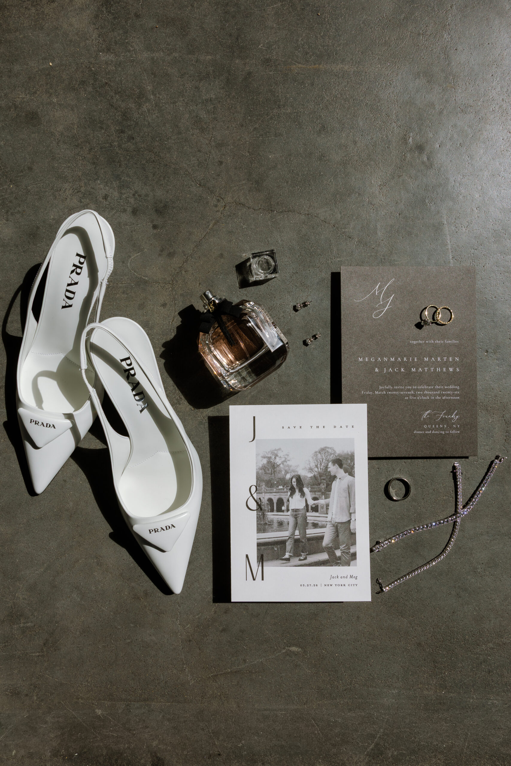 Flat lay photograph of white Prada high heels, wedding invitations, a 'Save the Date' card with a couple's photo, perfume bottle, rings, earrings, and a sparkling bracelet, arranged on a dark textured surface. Ideal for screen readers: this image showcases elegant wedding accessories and stationery, emphasizing luxury and romance.