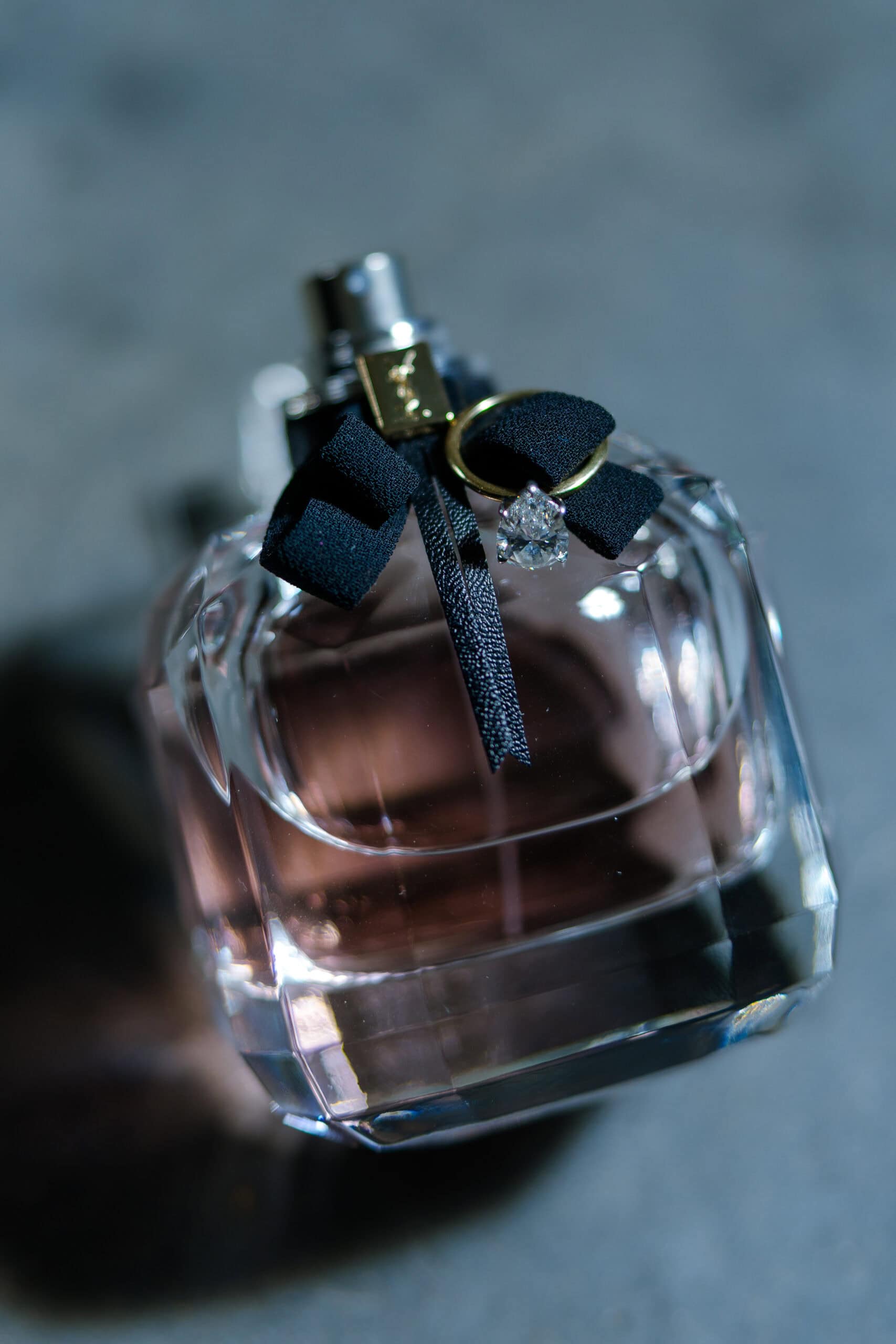 Close-up of a faceted perfume bottle with a black ribbon, a gold wedding band, and a pear-cut diamond ring styled on top at The Foundry.