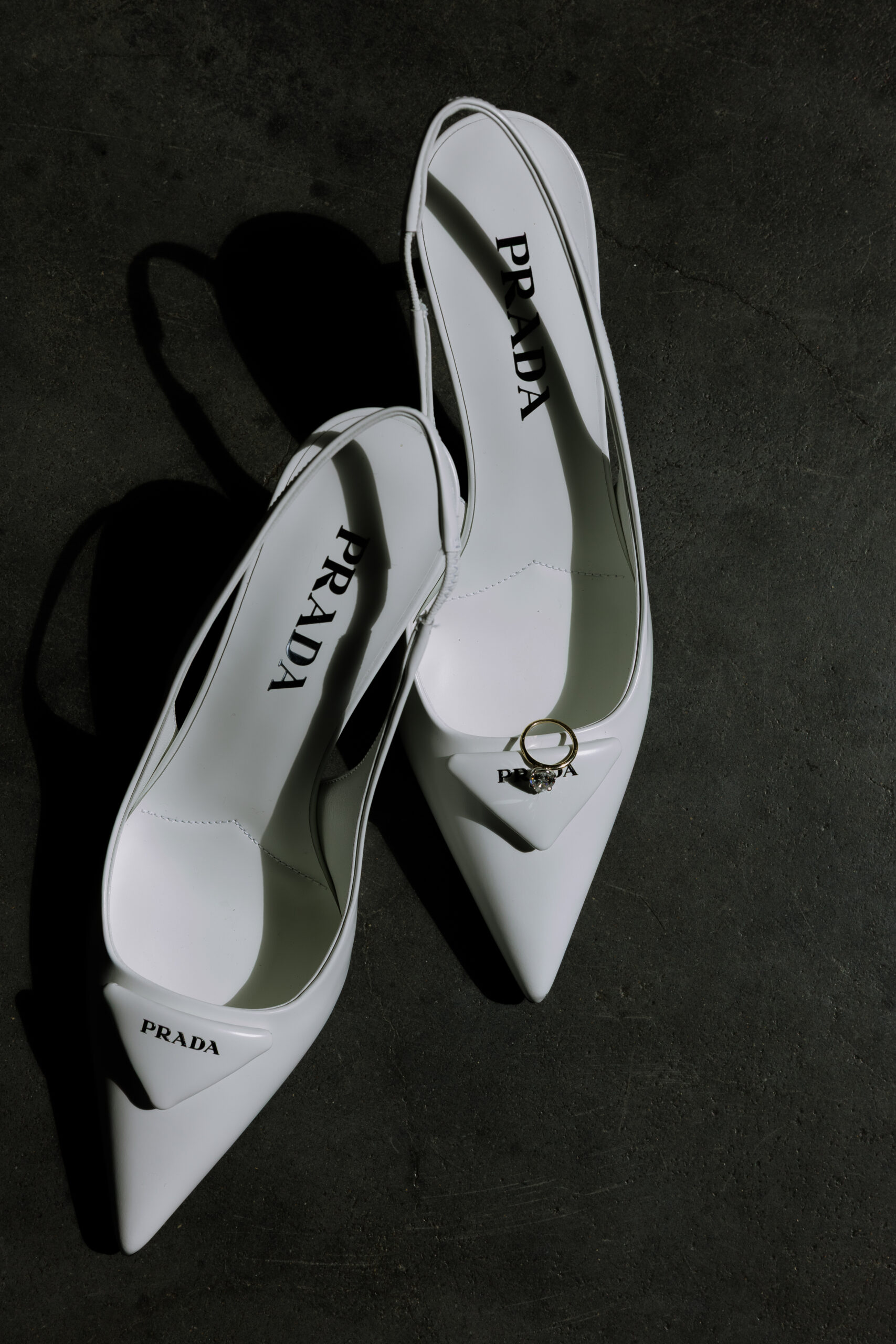 White Prada slingback heels on a dark stone floor with a diamond ring placed on one pointed toe, photographed as a wedding detail at The Foundry.