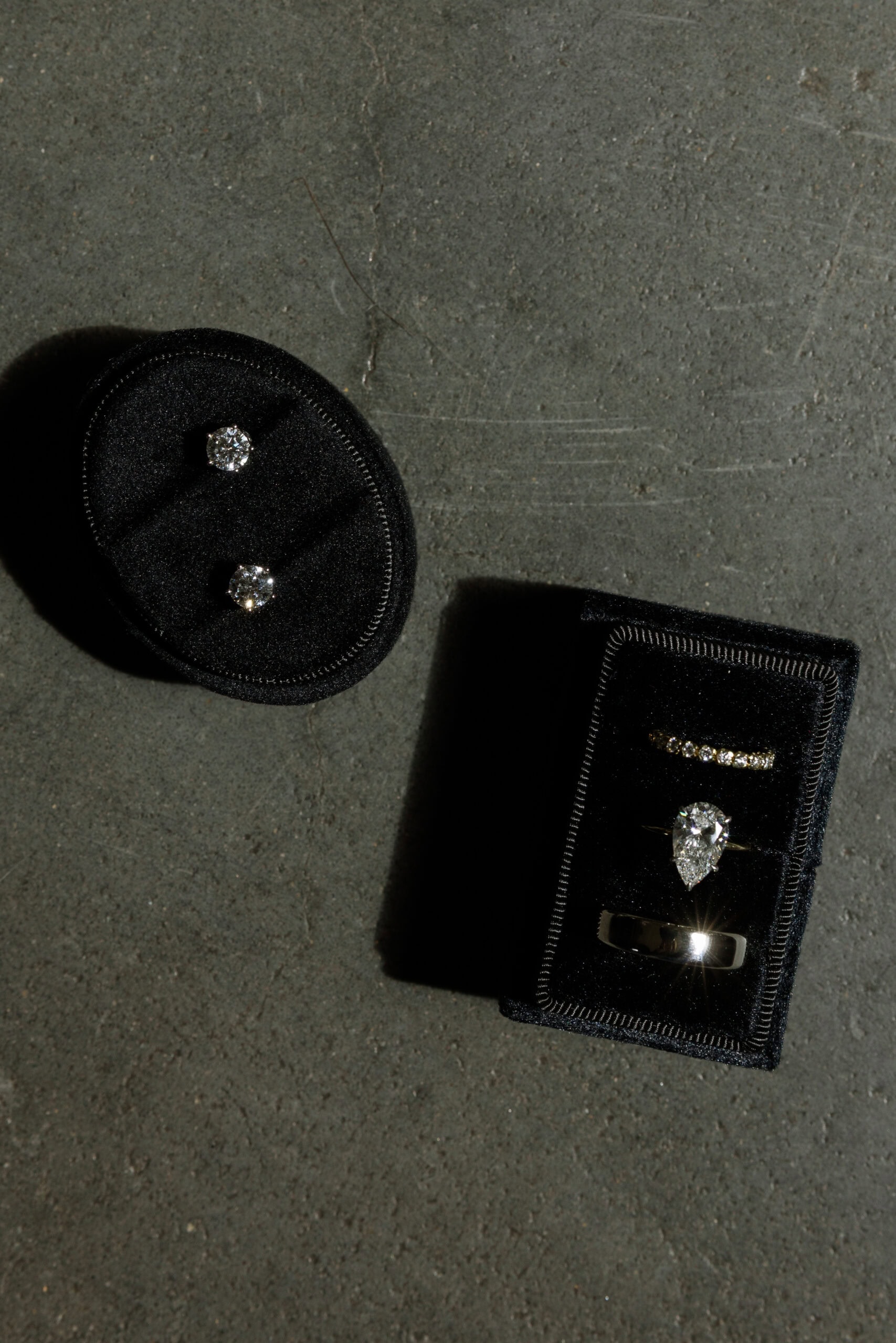 Flat lay of diamond stud earrings and wedding rings in black velvet boxes on a gray stone surface at The Foundry, photographed with dramatic light and shadow.