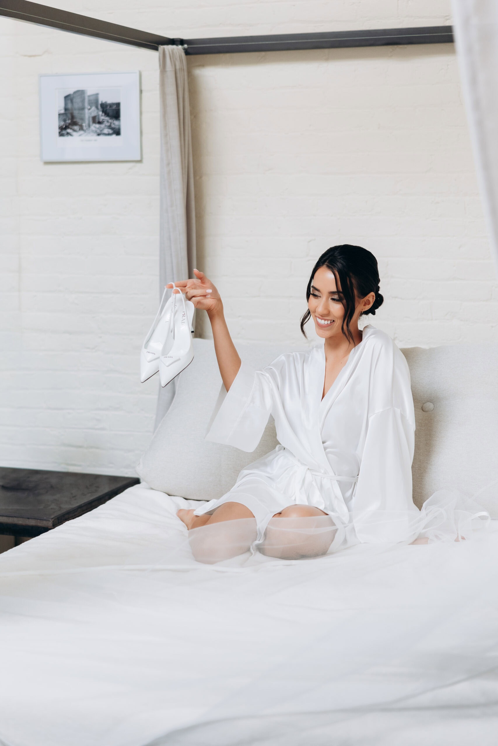 A bride in a white silk robe sits cross‑legged on a canopy bed in a bright room with a white brick wall, smiling as she holds up white Prada slingback heels; her tulle veil is spread across the bed. Photo taken March 27, 2026, during bridal prep.