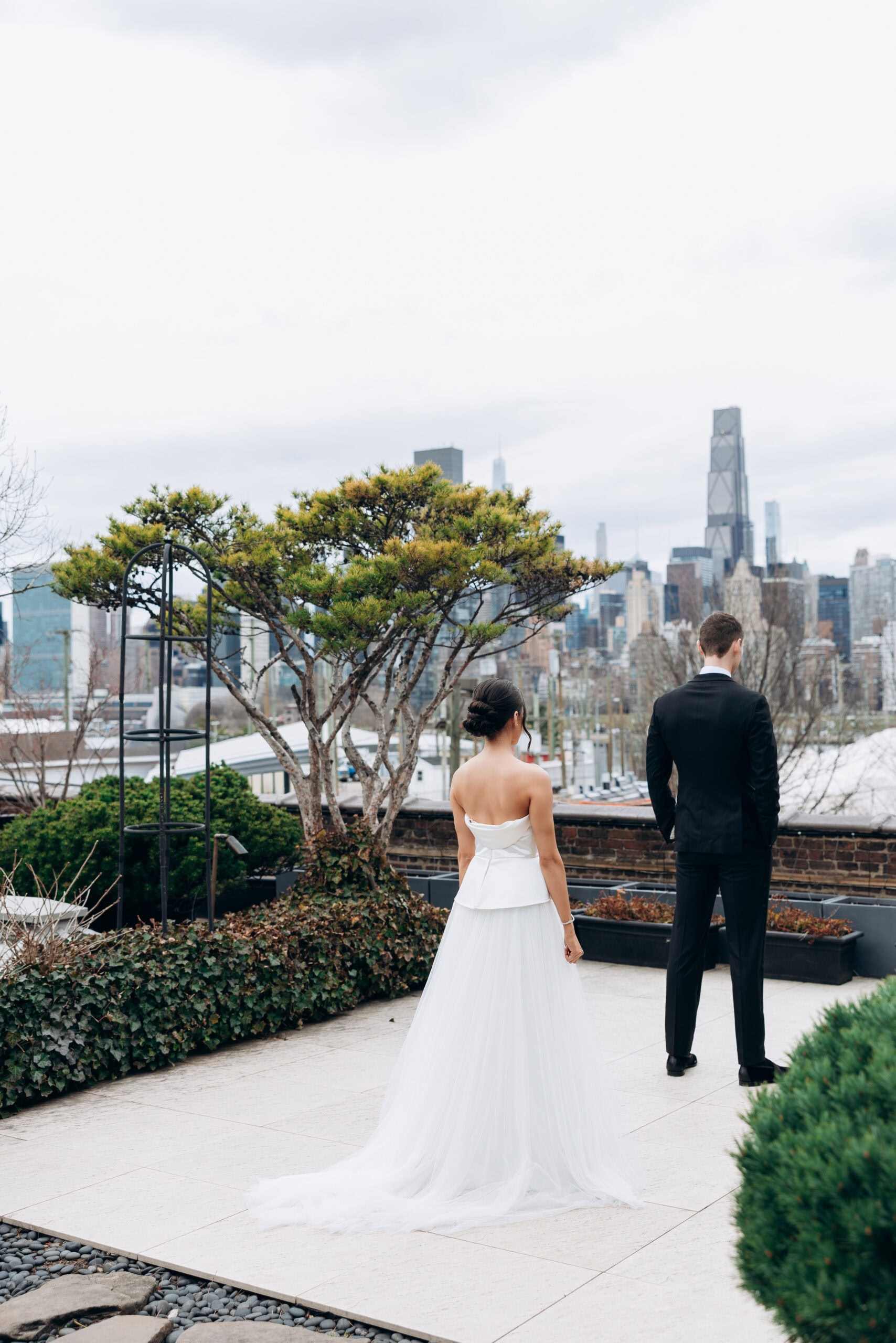 Rear view of a bride in a strapless white gown and a groom in a black suit standing on a terrace at The Foundry, facing the Manhattan skyline under an overcast sky.