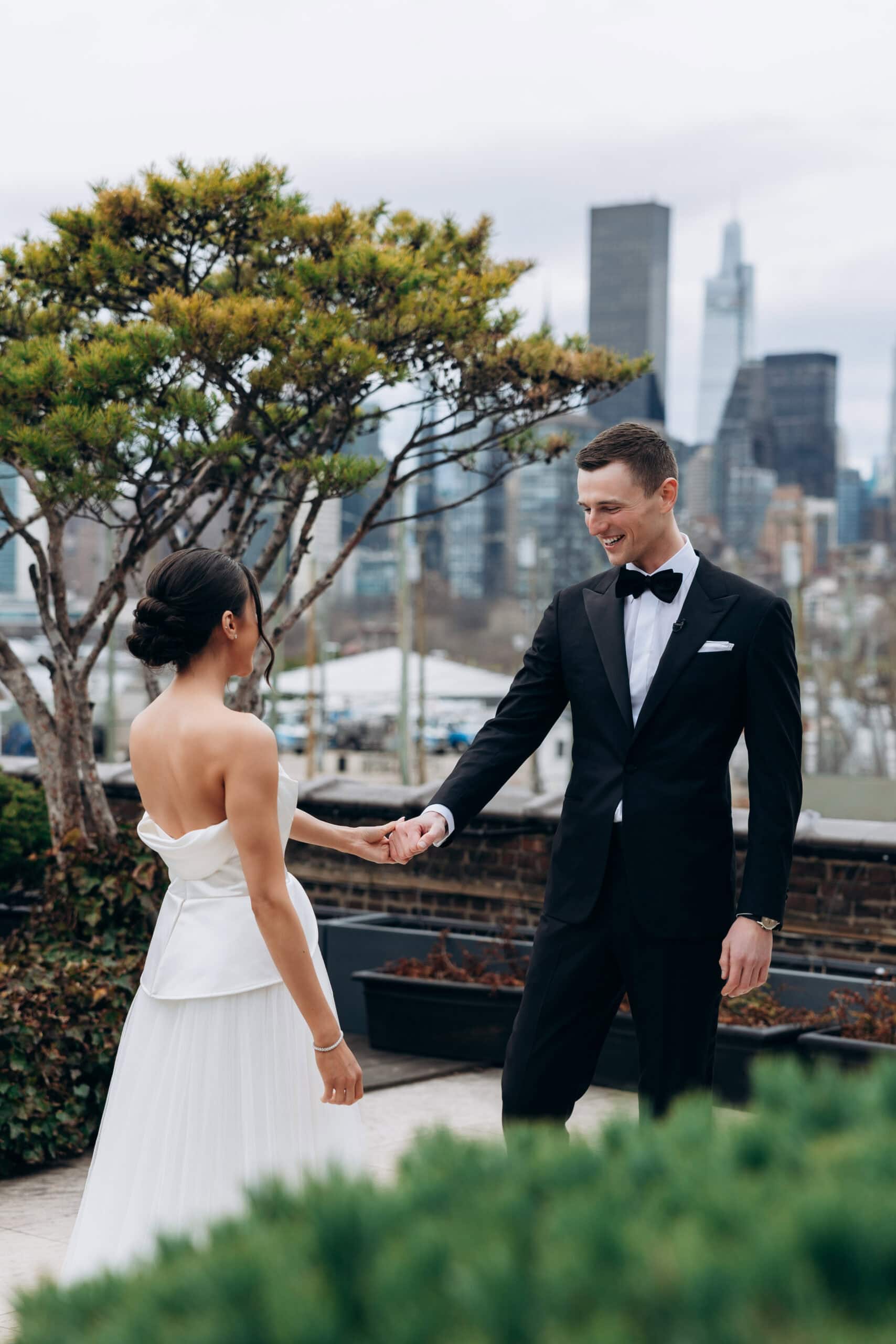 On March 27, 2026, a bride and groom share a first look on a Manhattan rooftop, holding hands beneath sculptural trees with the Midtown skyline behind.