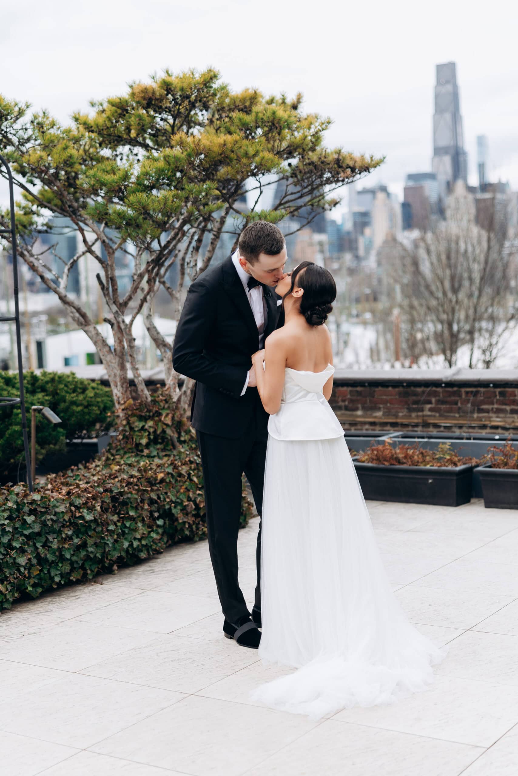 Bride and groom kiss on the terrace at The Foundry, with evergreen landscaping and a blurred Manhattan skyline in the background.