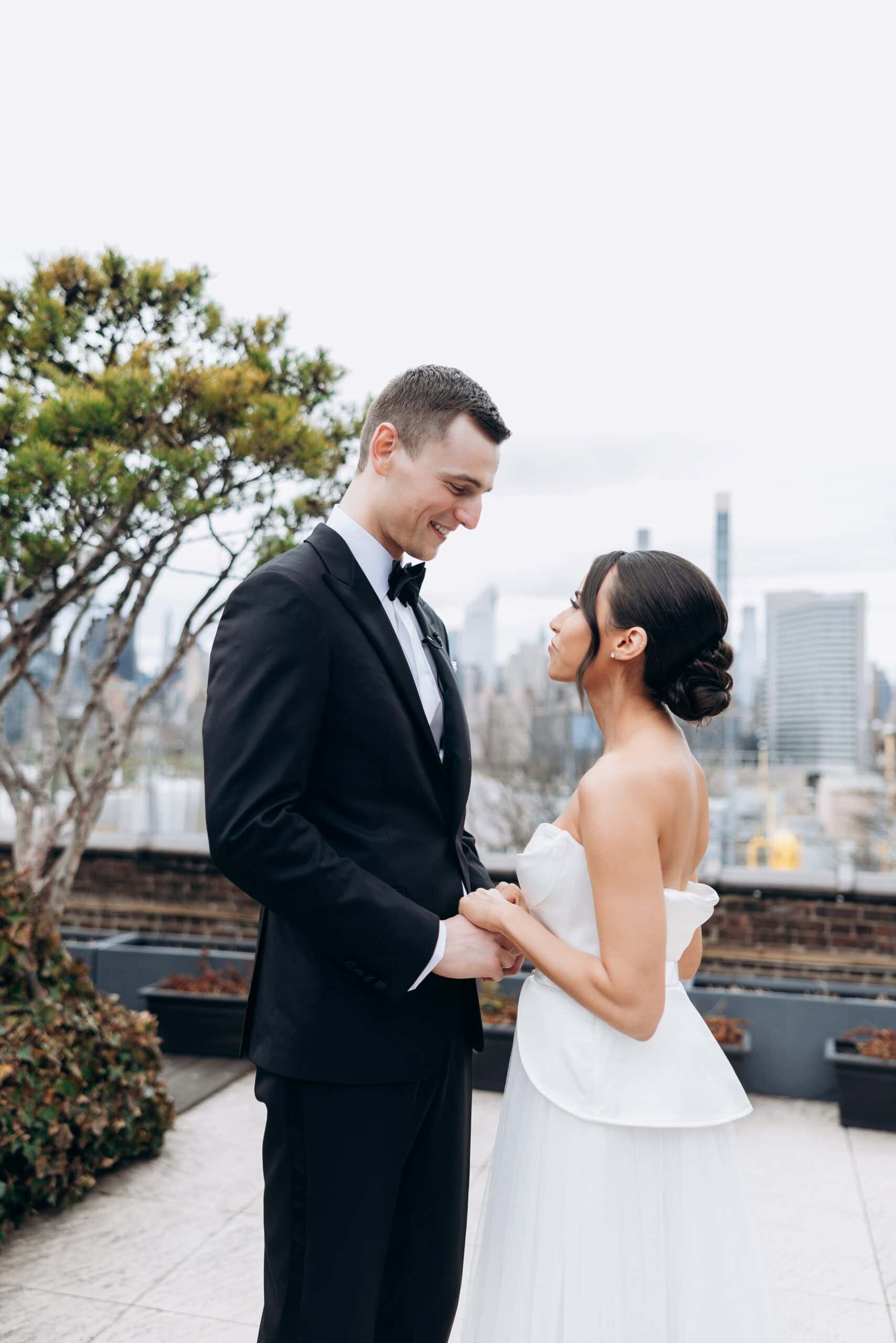 Bride and groom holding hands on a rooftop terrace at The Foundry, with the Manhattan skyline behind them; he wears a black tuxedo, she a strapless white gown.