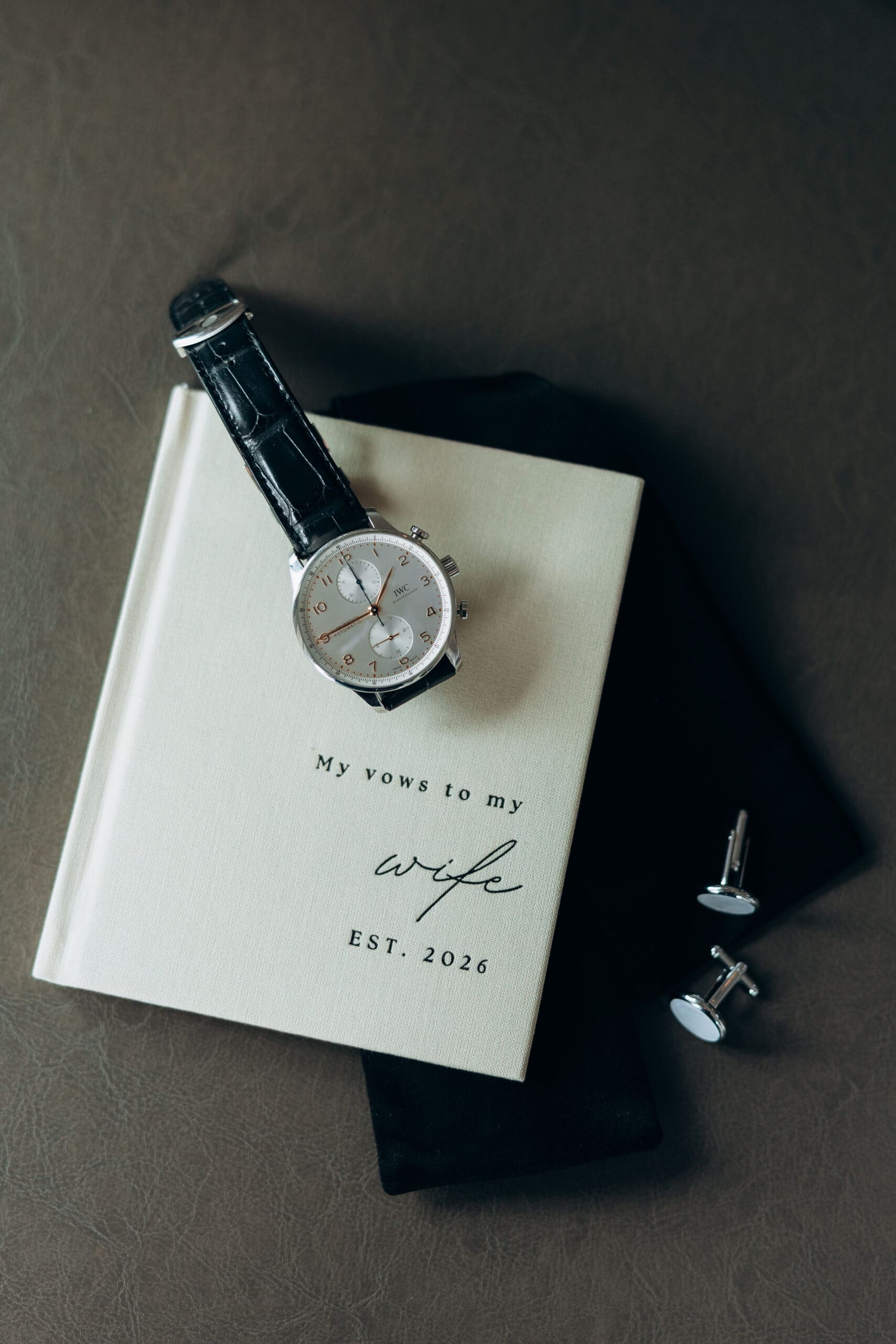 Flat lay of an IWC watch, cufflinks, dark fabric, and a vow book reading “My vows to my wife, Est. 2026” photographed at The Foundry.