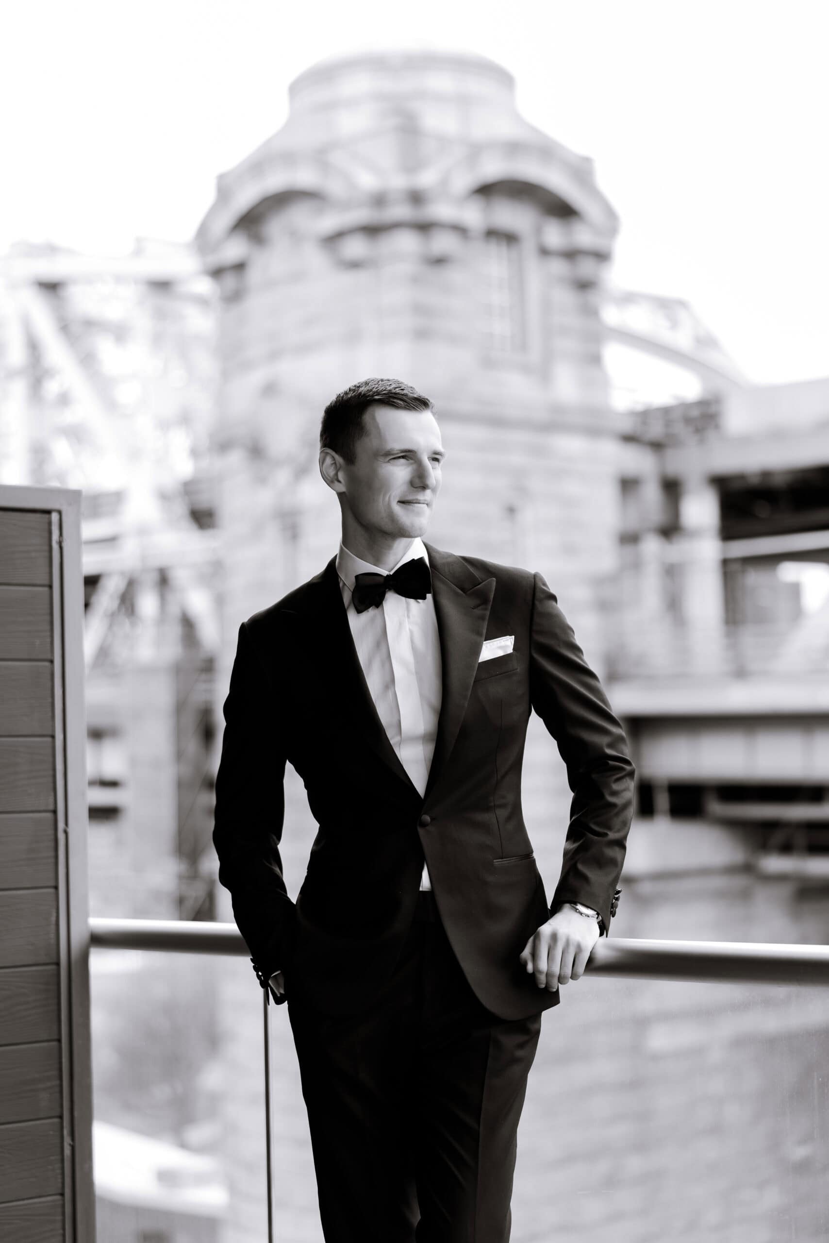 Black-and-white portrait of a man in a black tuxedo and bow tie leaning on a balcony railing at The Foundry, with blurred historic city architecture behind him.