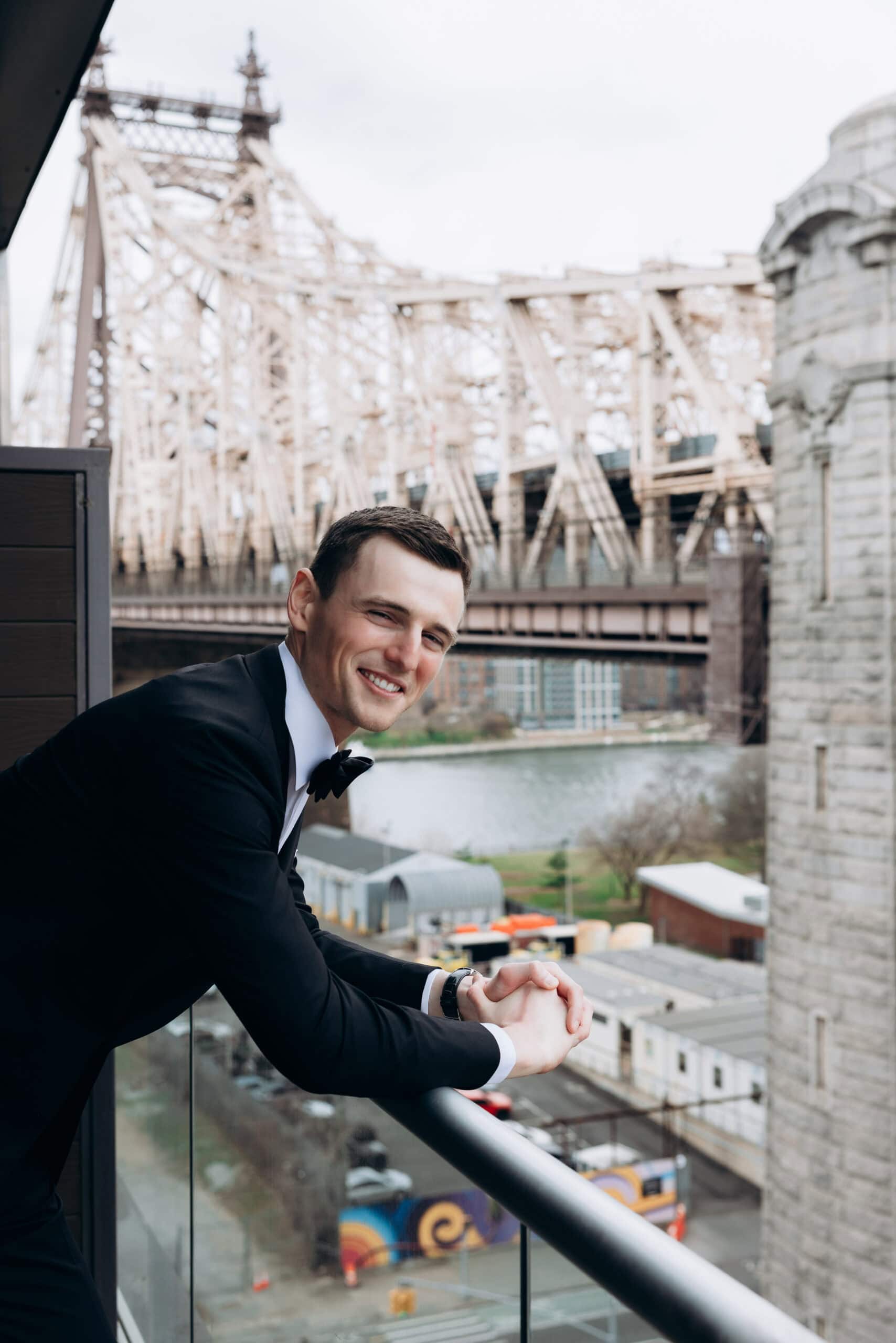 Man in a black tuxedo leaning on a balcony railing at The Foundry, with the Queensboro Bridge and waterfront visible behind him in soft focus.