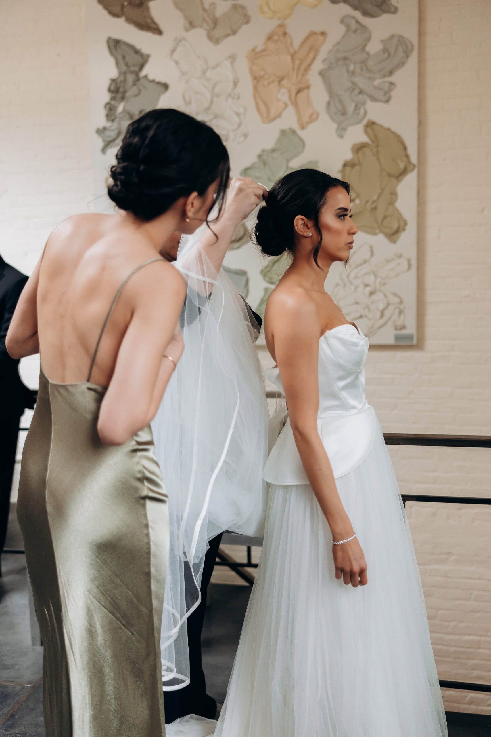 At The Foundry, a bridesmaid adjusts a bride’s veil indoors as the bride stands in profile wearing a satin and tulle gown before an abstract wall artwork.