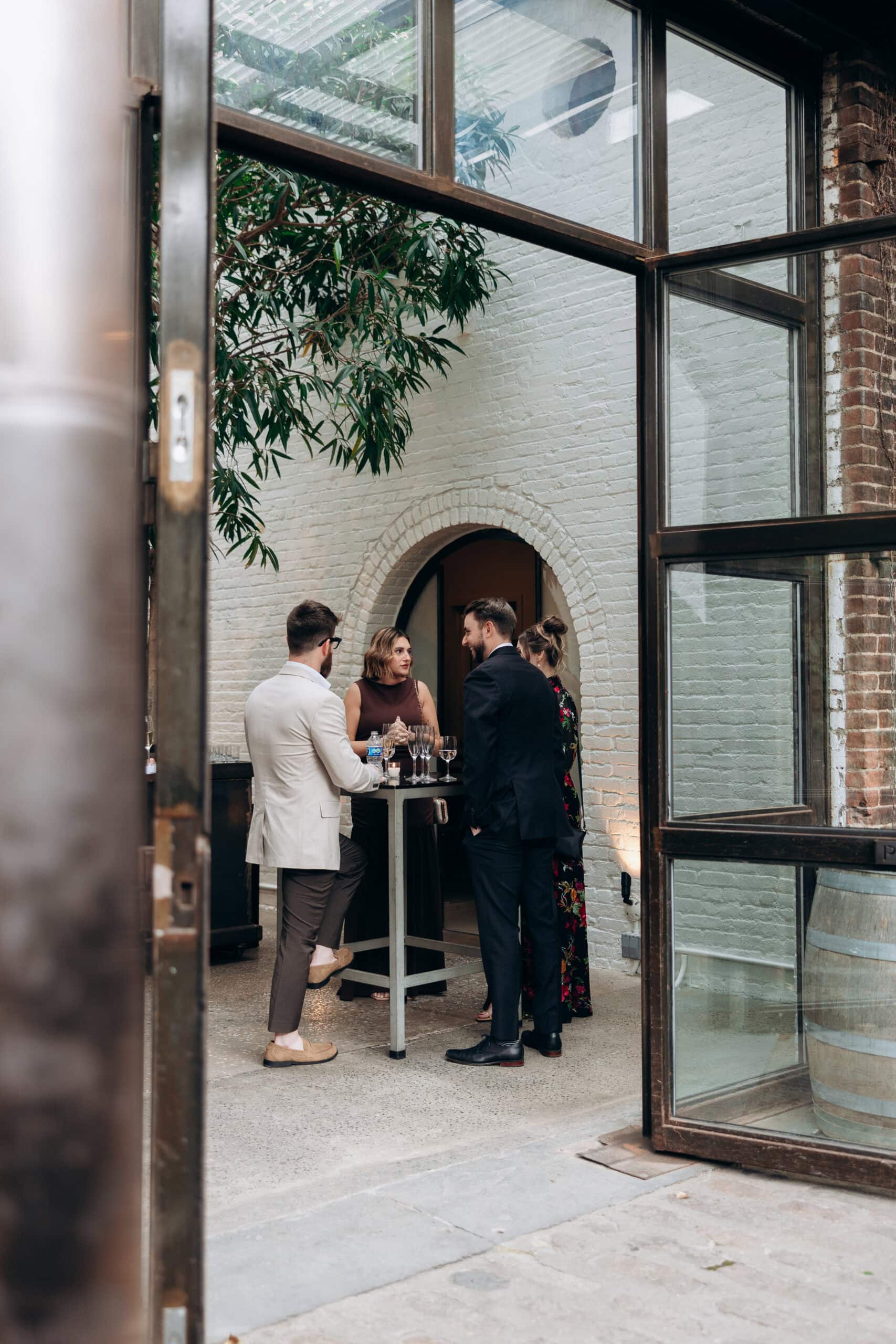 Four wedding guests stand around a high-top table during cocktail hour at The Foundry, seen through open steel-framed doors in a brick courtyard space.