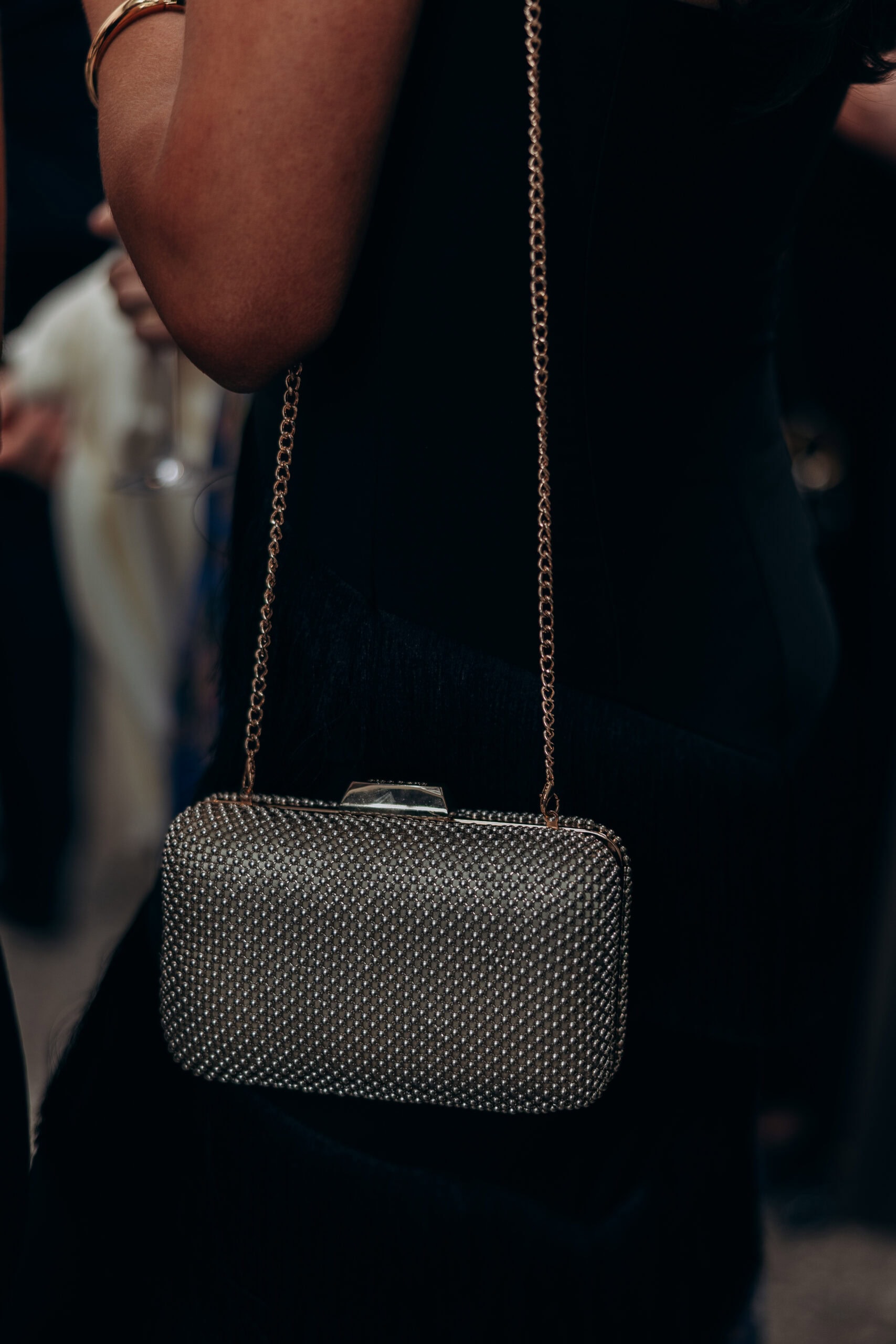 Close-up of a wedding guest at The Foundry wearing a black dress and gold bangle, carrying a crystal-studded clutch with chain strap in dim reception light.