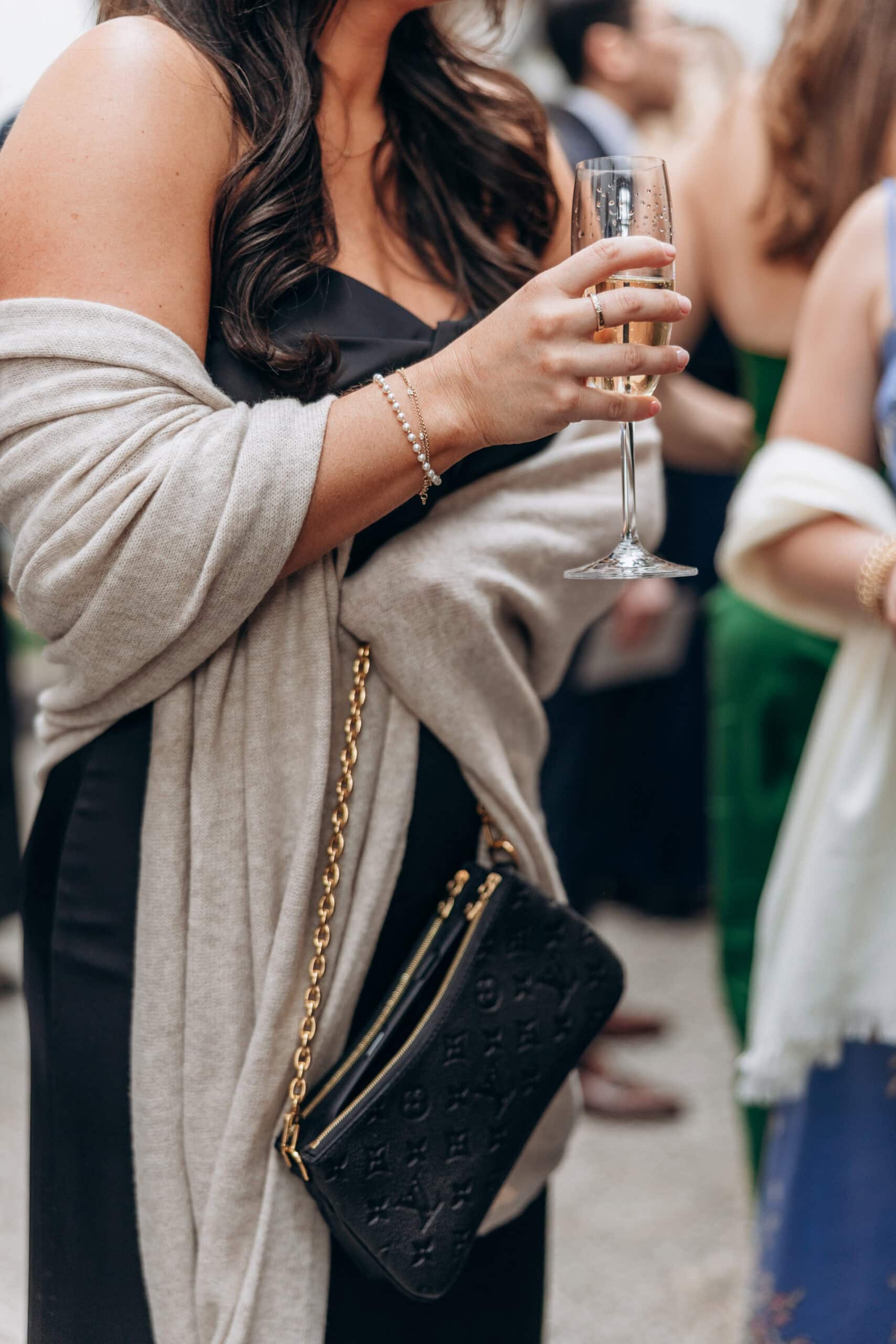 Close-up of a wedding guest at The Foundry holding a champagne flute, wearing a black dress, cream shawl, pearl bracelet, and black embossed chain bag.