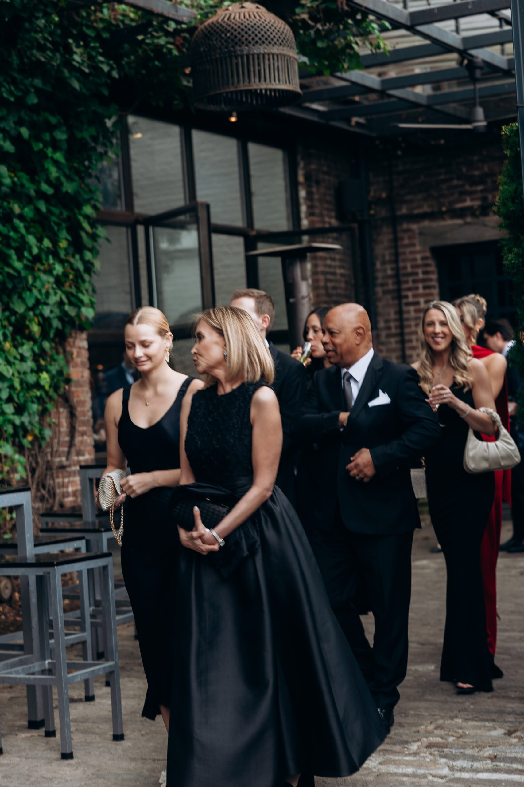 Wedding guests in black-tie attire walk through the ivy-lined courtyard at The Foundry, with brick walls, a glass canopy, and cocktail-hour ambiance.