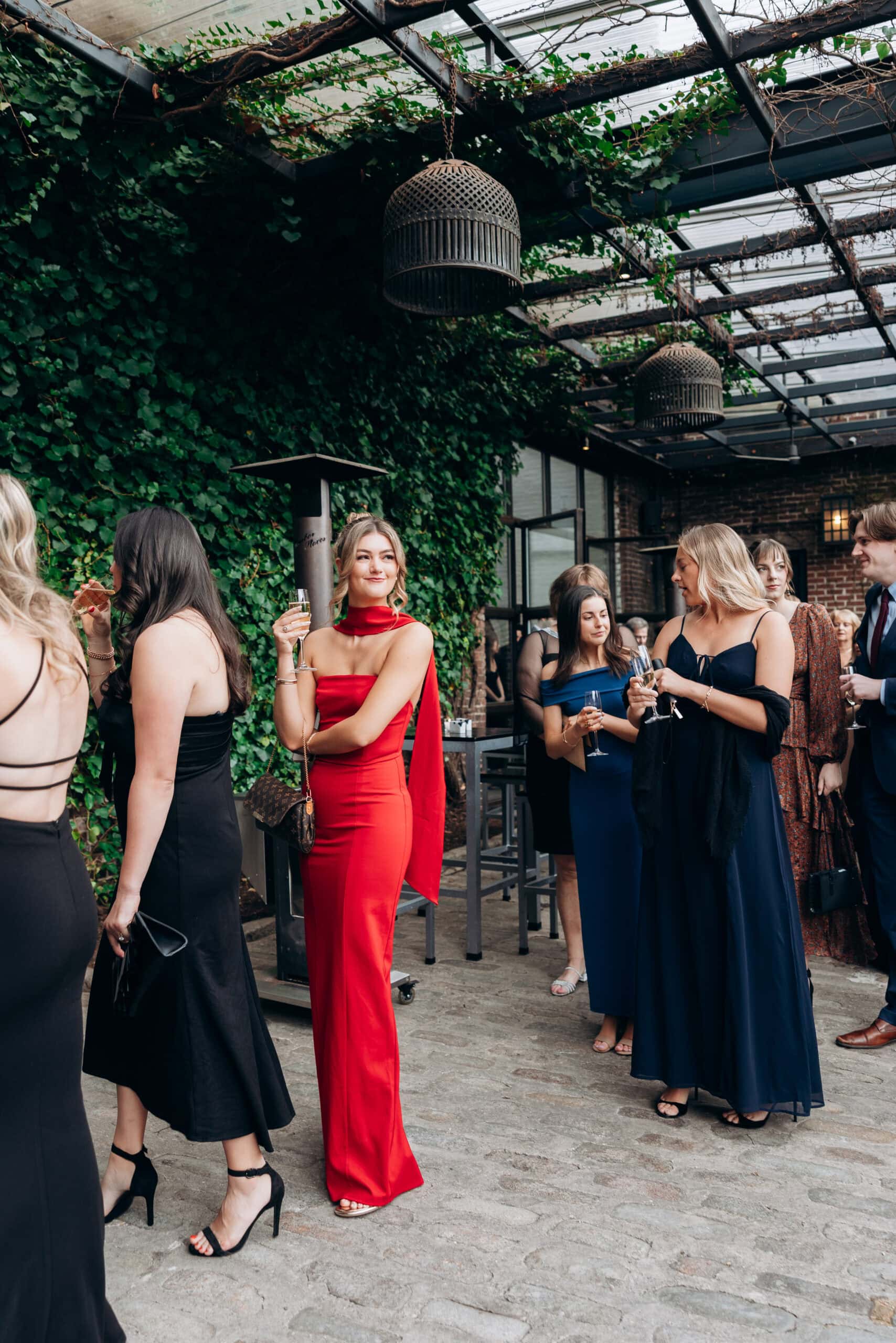 Guests in formal eveningwear mingle with champagne during a wedding cocktail hour in the ivy-covered courtyard at The Foundry, beneath a glass canopy and hanging lanterns.