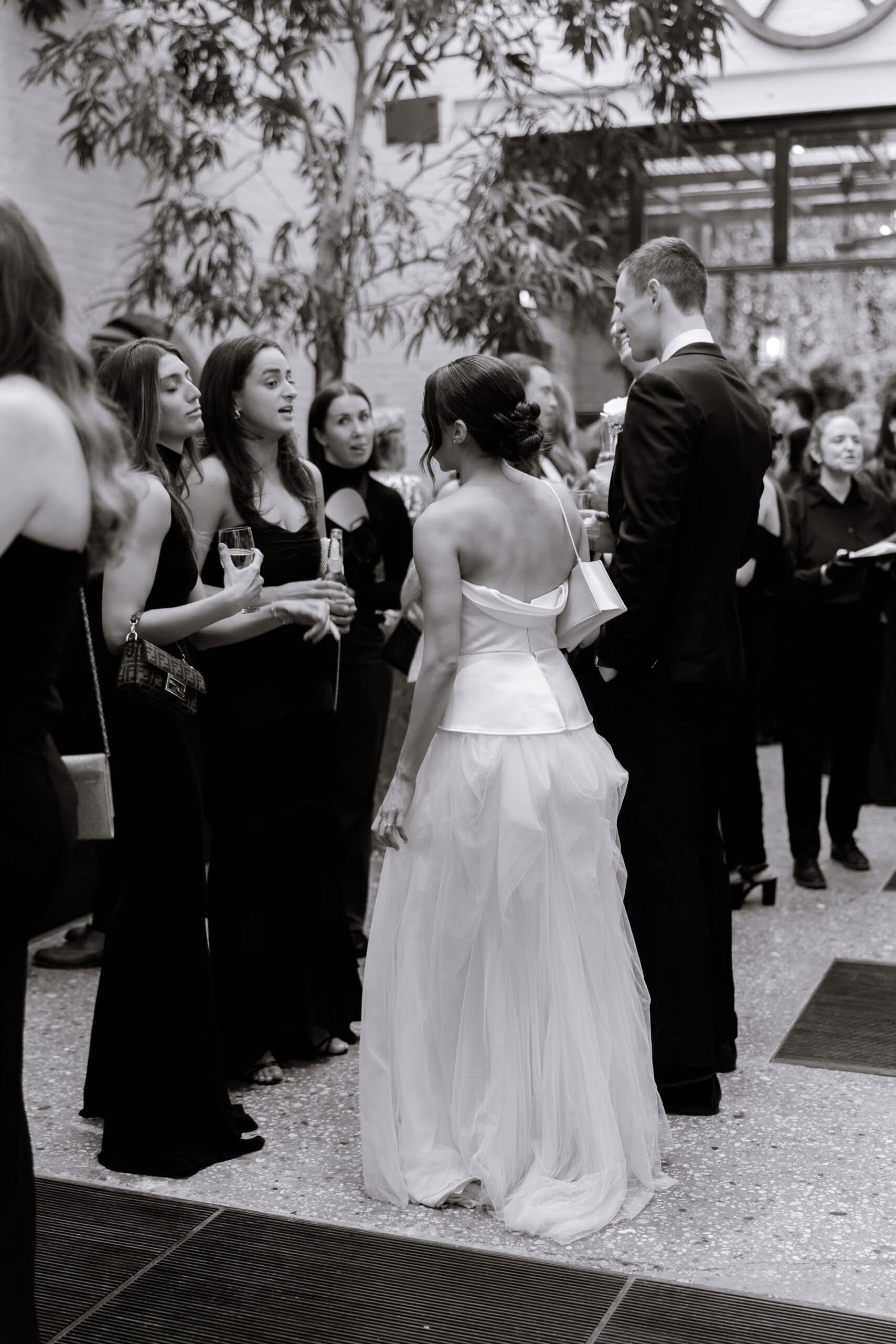 Black-and-white photo of a bride in a backless satin-and-tulle gown speaking with guests during cocktail hour at The Foundry, in a bright indoor garden venue.