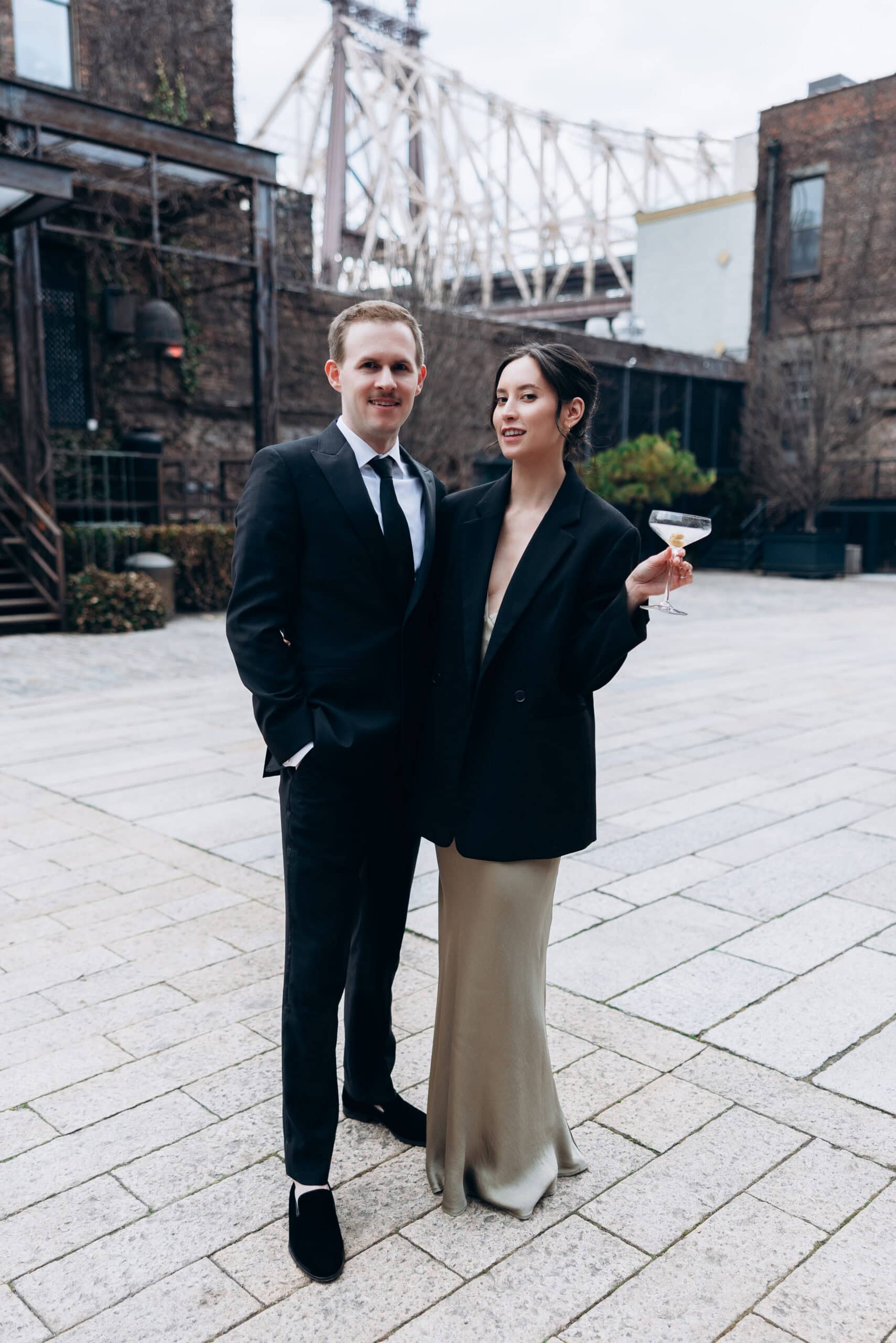 Two guests in formal black attire pose in a stone courtyard at The Foundry, with brick buildings and the Queensboro Bridge behind them; one holds a coupe glass.